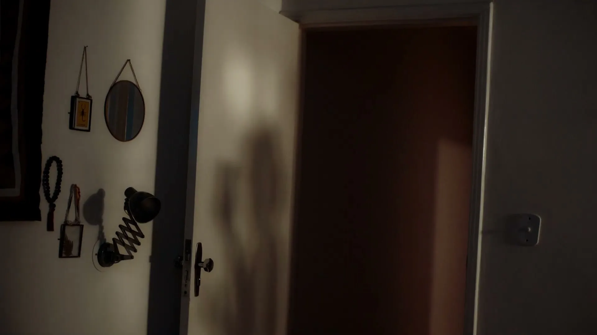 A dark room with a partially open door reveals a faint shadow of a person passing by, creating a mysterious atmosphere.