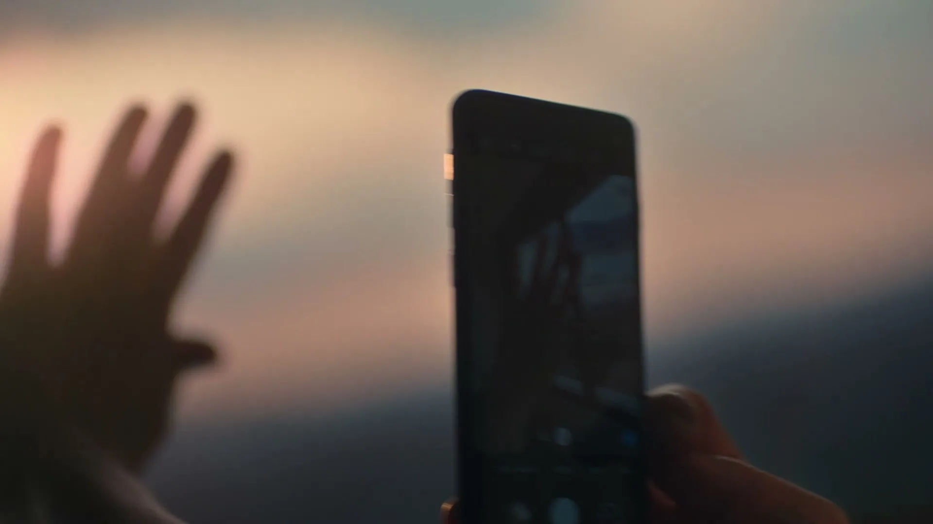 A person with glasses holds a smartphone to record a hand waving against a blurred sunset sky.