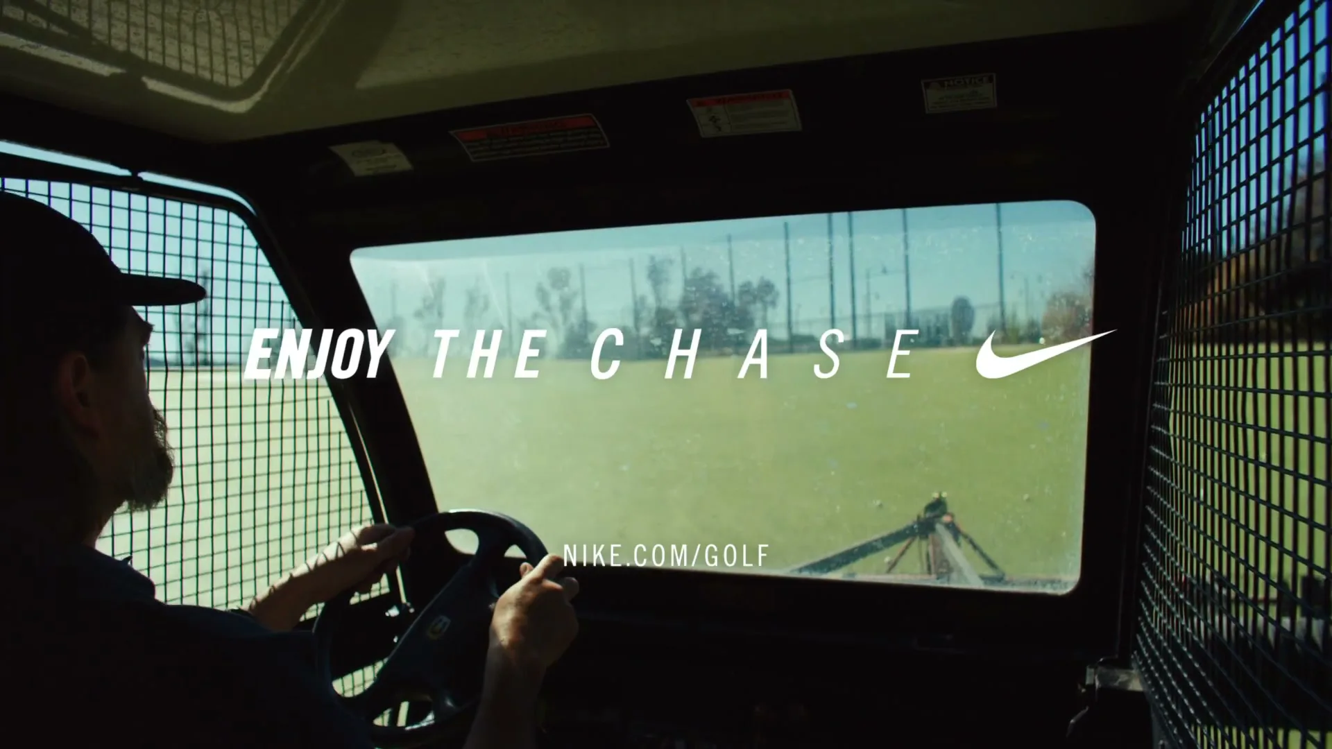 A man with a beard and a cap drives a utility vehicle across a golf course, with text overlayed on the screen.