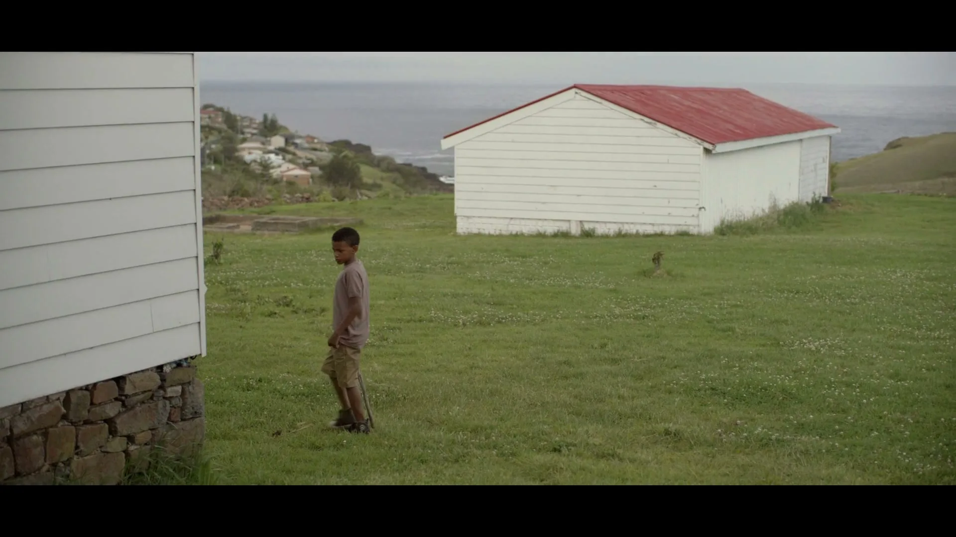 A young boy in a brown t-shirt and shorts walks with a stick in a grassy field next to a white building with a red roof, overlooking the ocean.