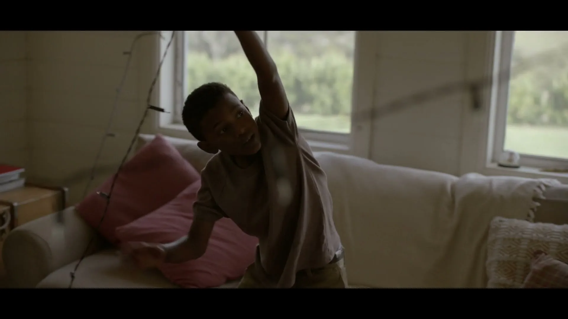 A young boy in a t-shirt and shorts stretches his arm up, holding a string of lights in a cozy living room.