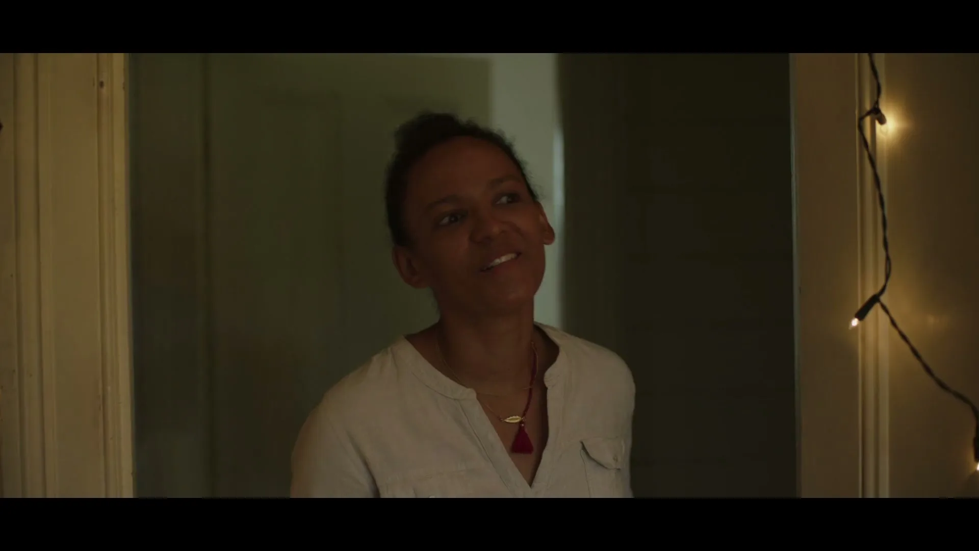 A woman with dark skin and hair tied up, wearing a light shirt and necklace, smiles while looking up and to the right in a dimly lit room with string lights.