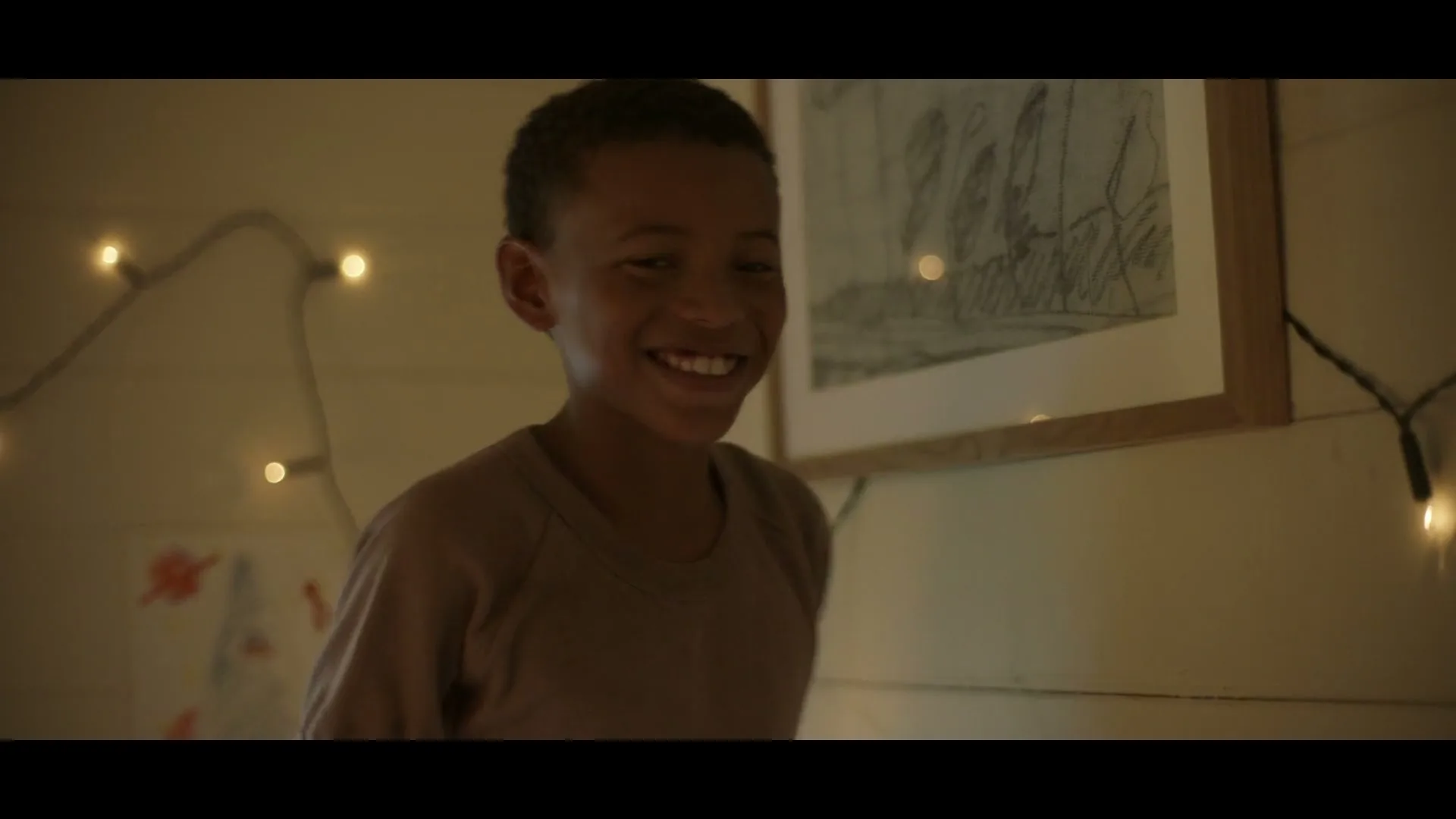 A young boy with short dark hair, wearing a brown t-shirt, smiles broadly while standing next to a wall adorned with string lights and framed drawings.