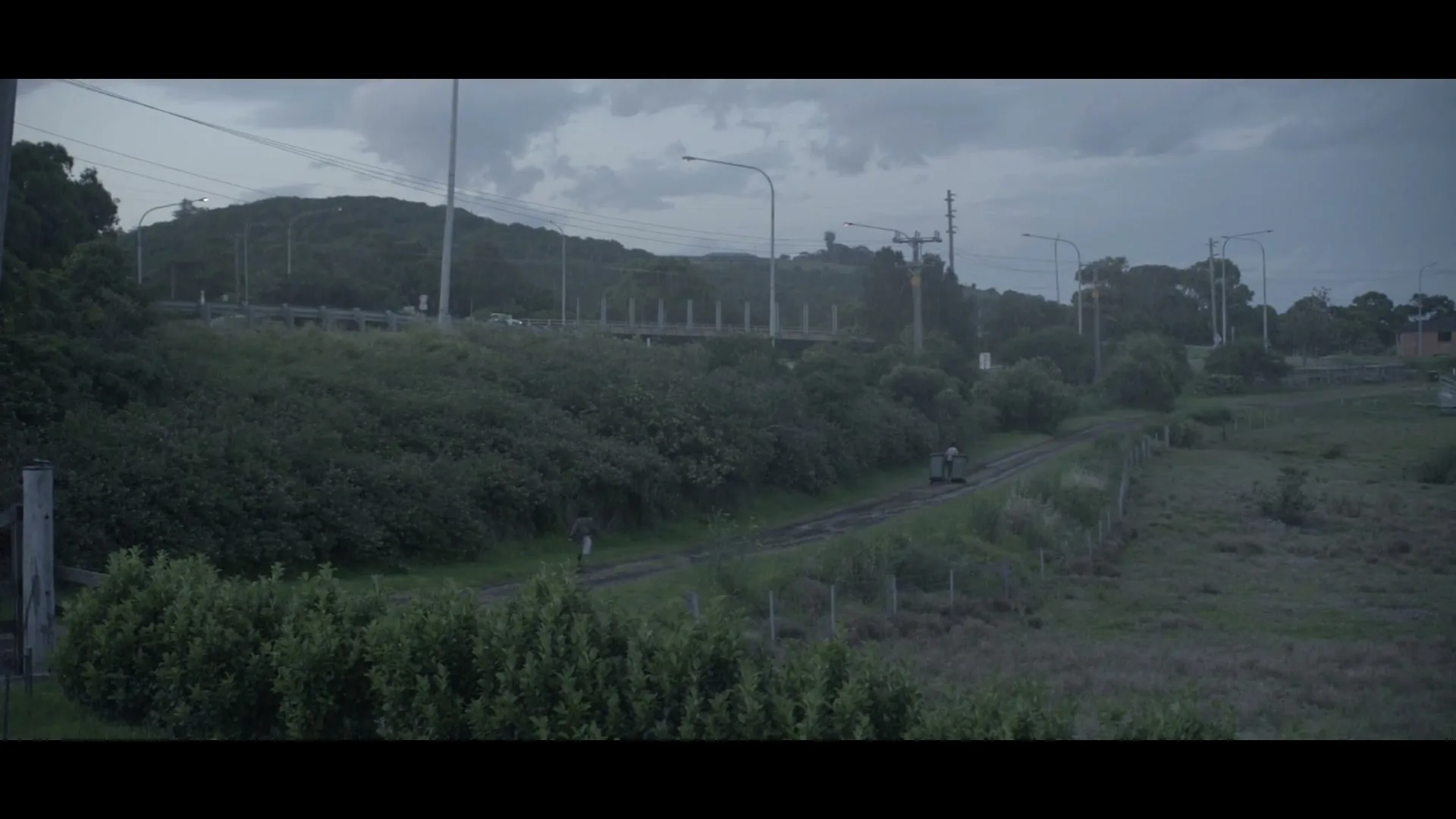 Two people walk along a dirt path next to train tracks, with a grassy hill and highway in the background under a cloudy sky.