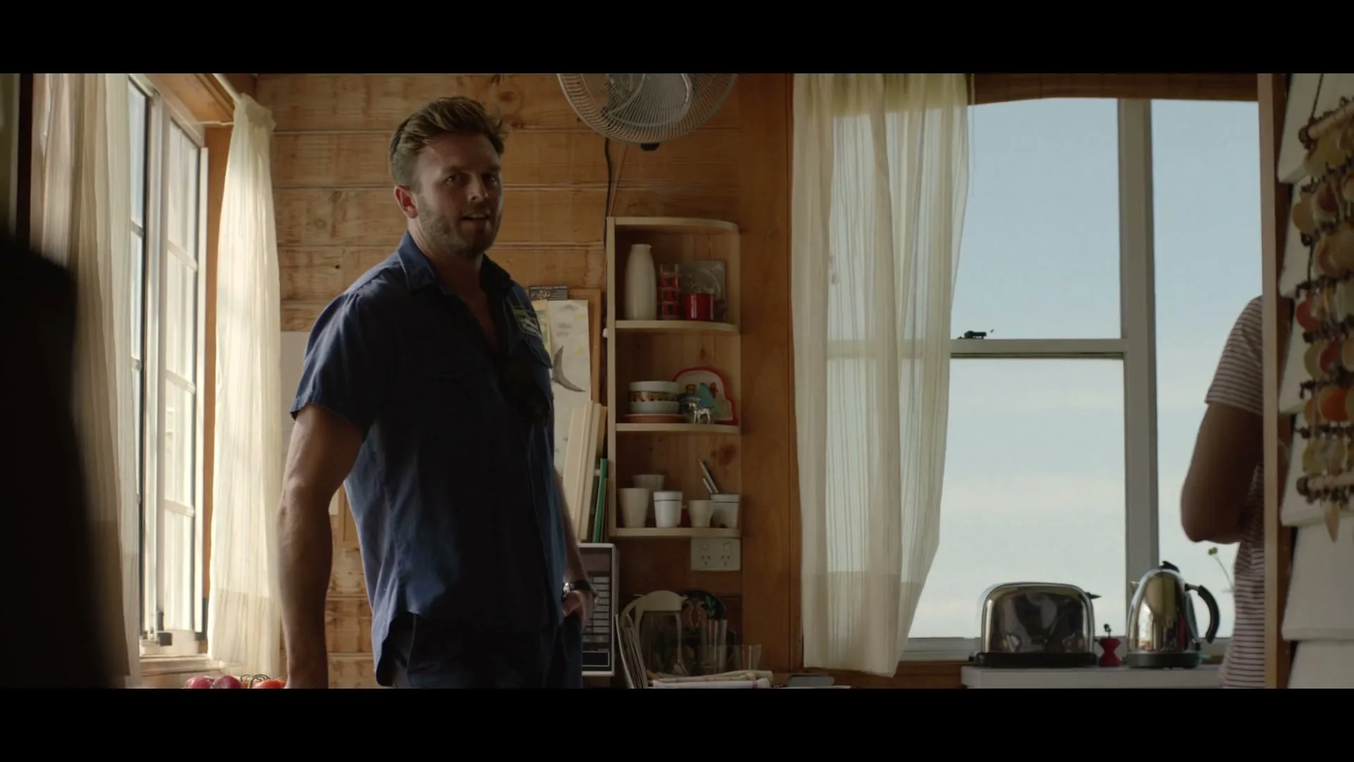 A man in a blue shirt stands in a rustic kitchen, looking towards a woman who is partially visible on the right, gesturing with her hand.