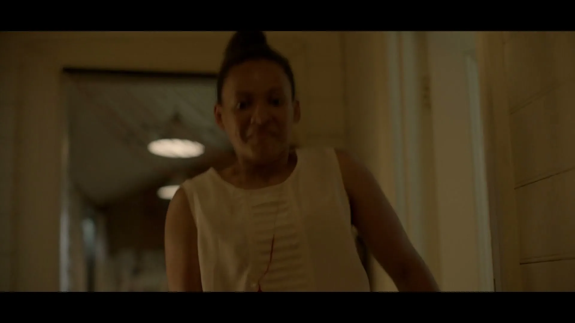 A woman with dark skin and hair tied up, wearing a white top, looks shocked and then confused in a dimly lit hallway.