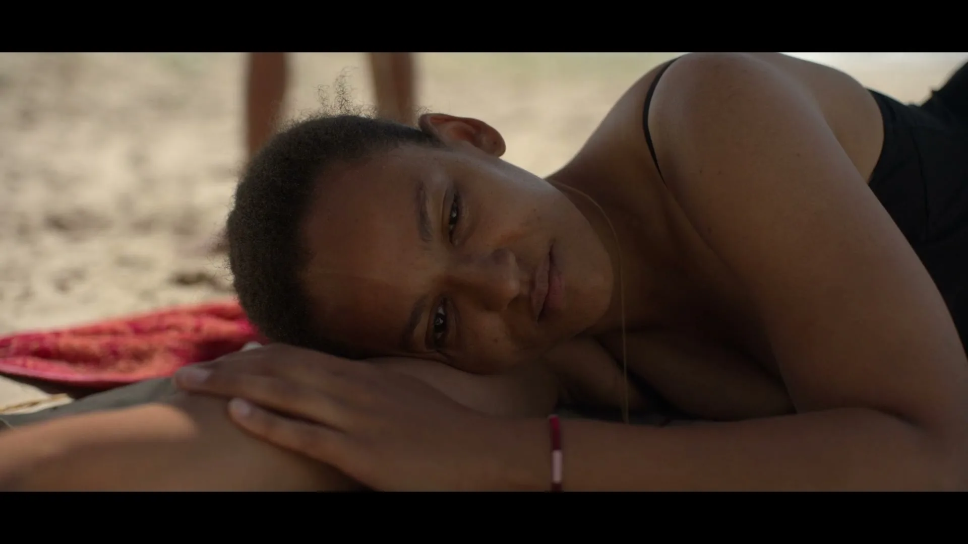 A young woman with dark skin and hair, wearing a black top, lies on a beach, resting her head on her arm, and slowly opens her eyes.