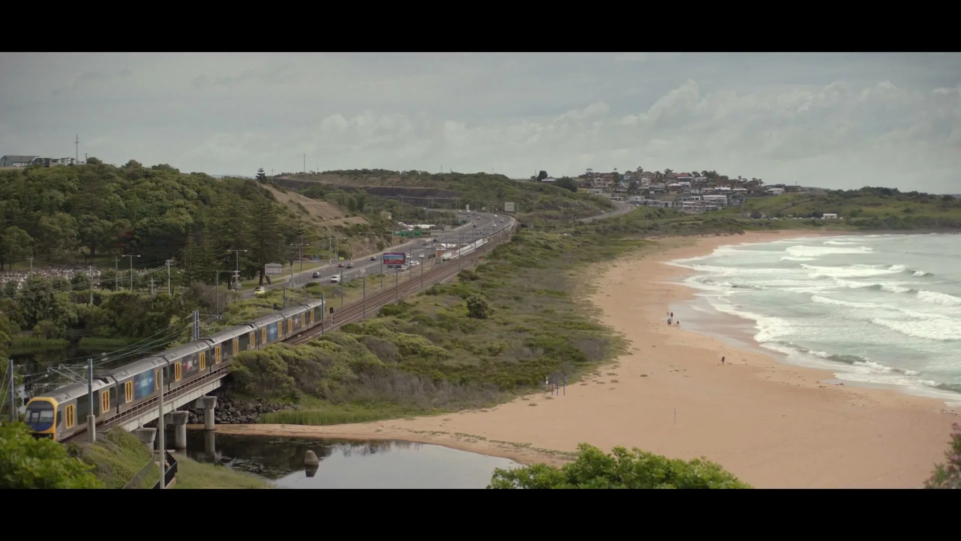A long shot of a train crossing a bridge next to a highway and a beach with waves crashing, under a cloudy sky.