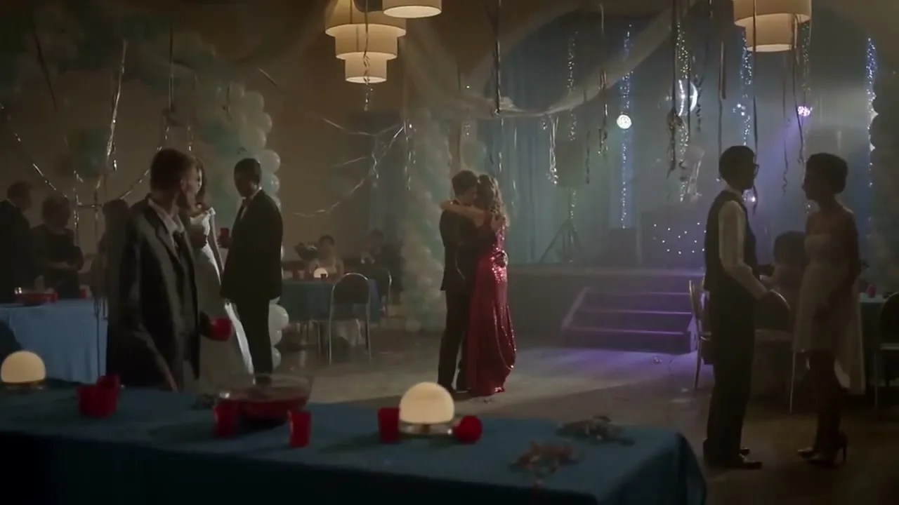 A young couple slow dances in the middle of a high school prom, surrounded by other students and decorations.