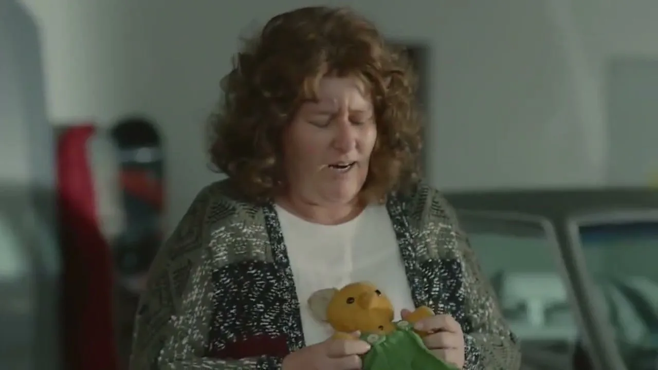 A middle-aged woman with curly brown hair, wearing a patterned cardigan, holds a teddy bear and looks down with a pained expression in a garage.