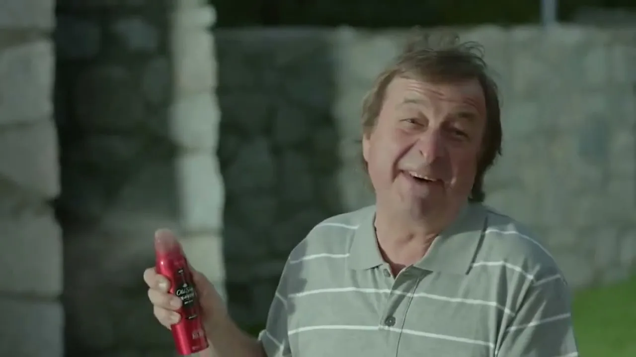 A middle-aged man with messy hair, wearing a striped polo shirt, holds a red Old Spice deodorant can and sprays it while smiling at the camera in an outdoor setting.