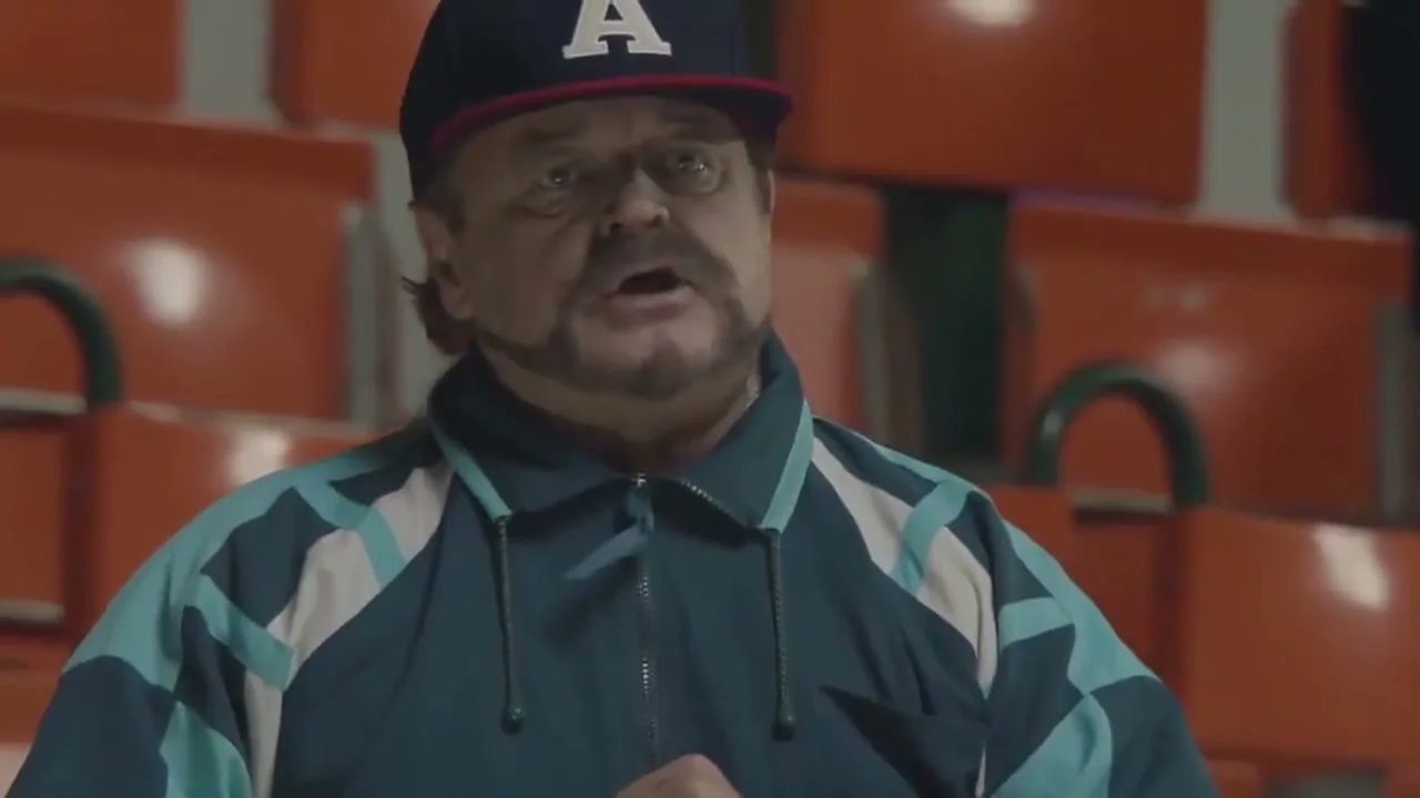 A middle-aged man with a mustache and a baseball cap sings in a stadium, looking concerned.