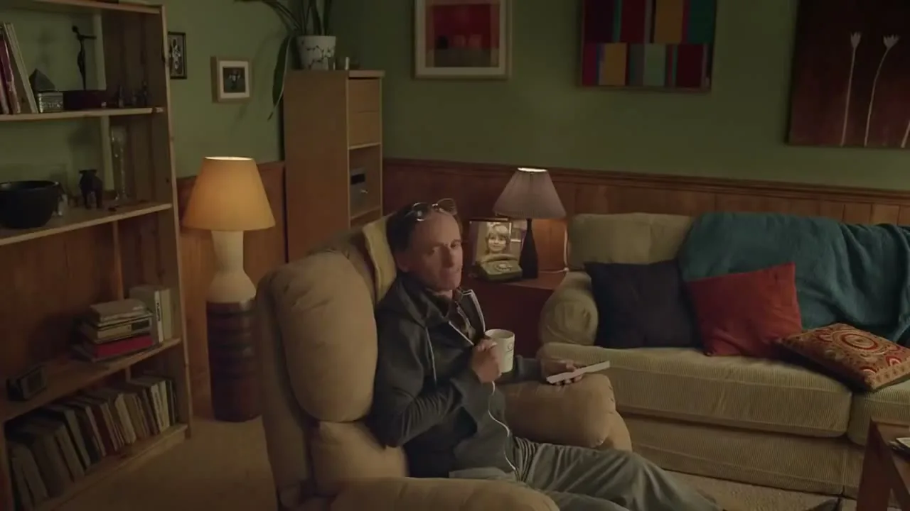 A middle-aged man in a grey tracksuit sits in a recliner, holding a mug and remote, looking at a framed photo of a child in a cozy living room.