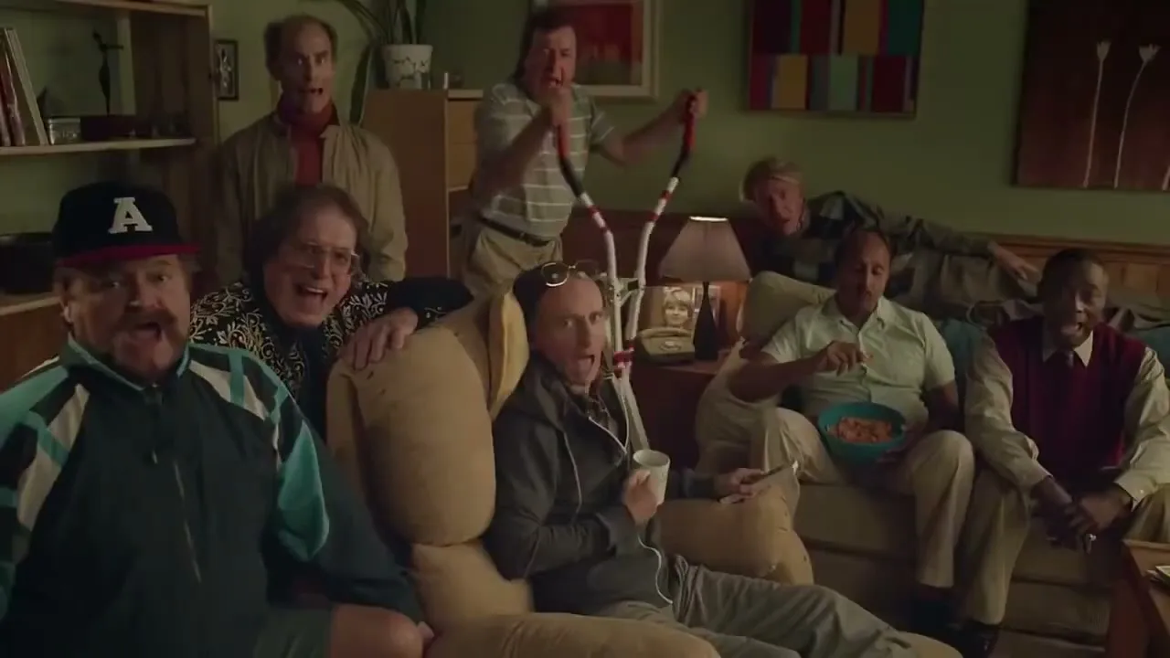 A group of diverse men, mostly middle-aged, are gathered in a living room, some sitting on a couch, some standing, all looking towards the camera with open mouths as if singing.