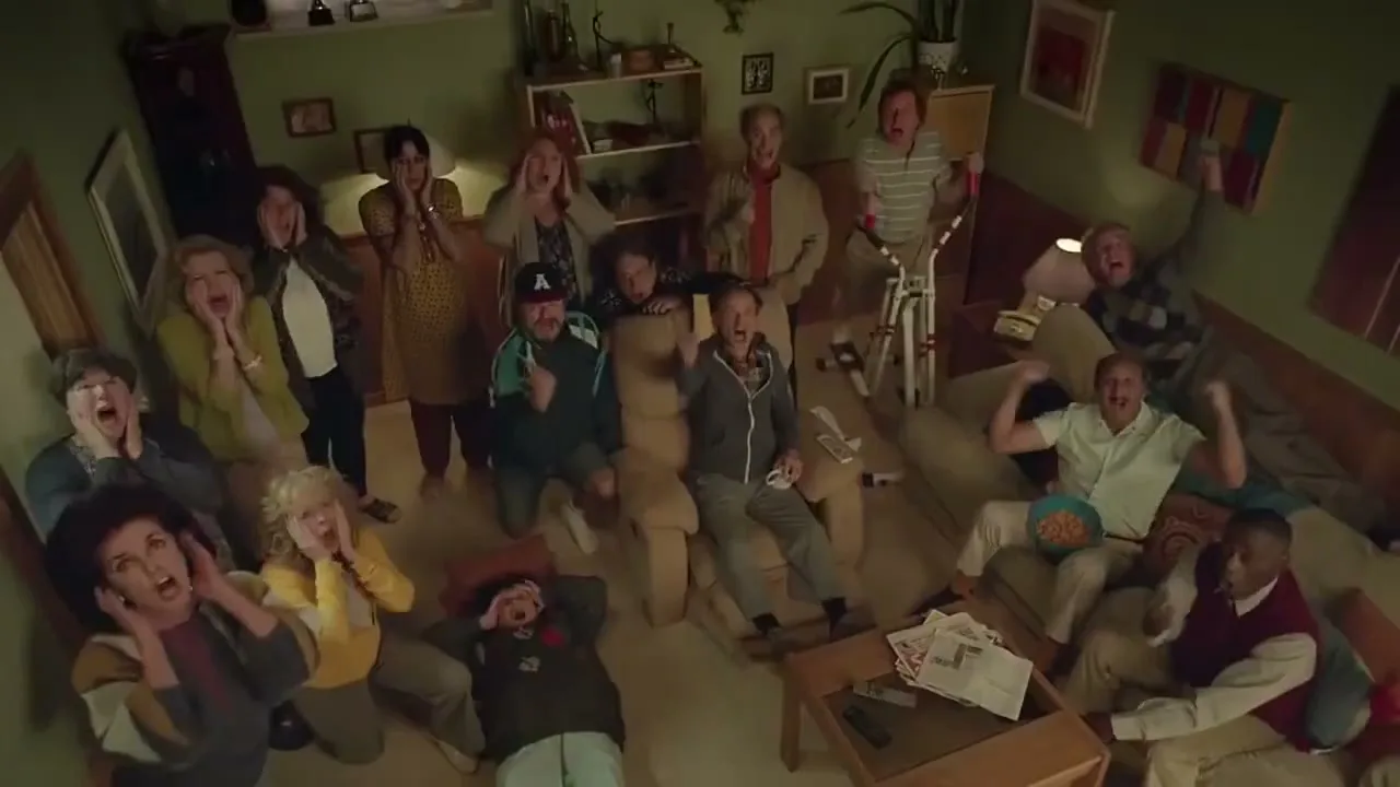 A large group of diverse people, including men and women of various ages, are gathered in a living room, all looking up with expressions of shock and awe.