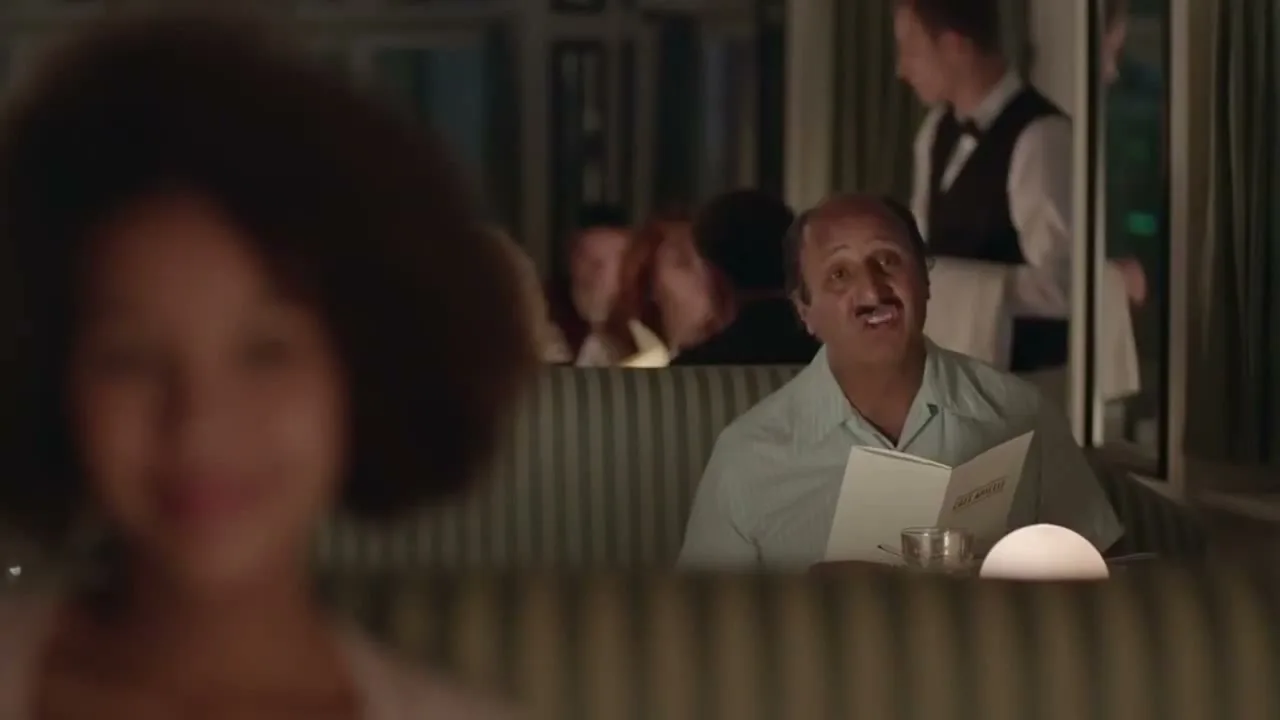 A man with a mustache in a light blue shirt holds a menu and sings in a restaurant booth, with a blurred woman in the foreground.