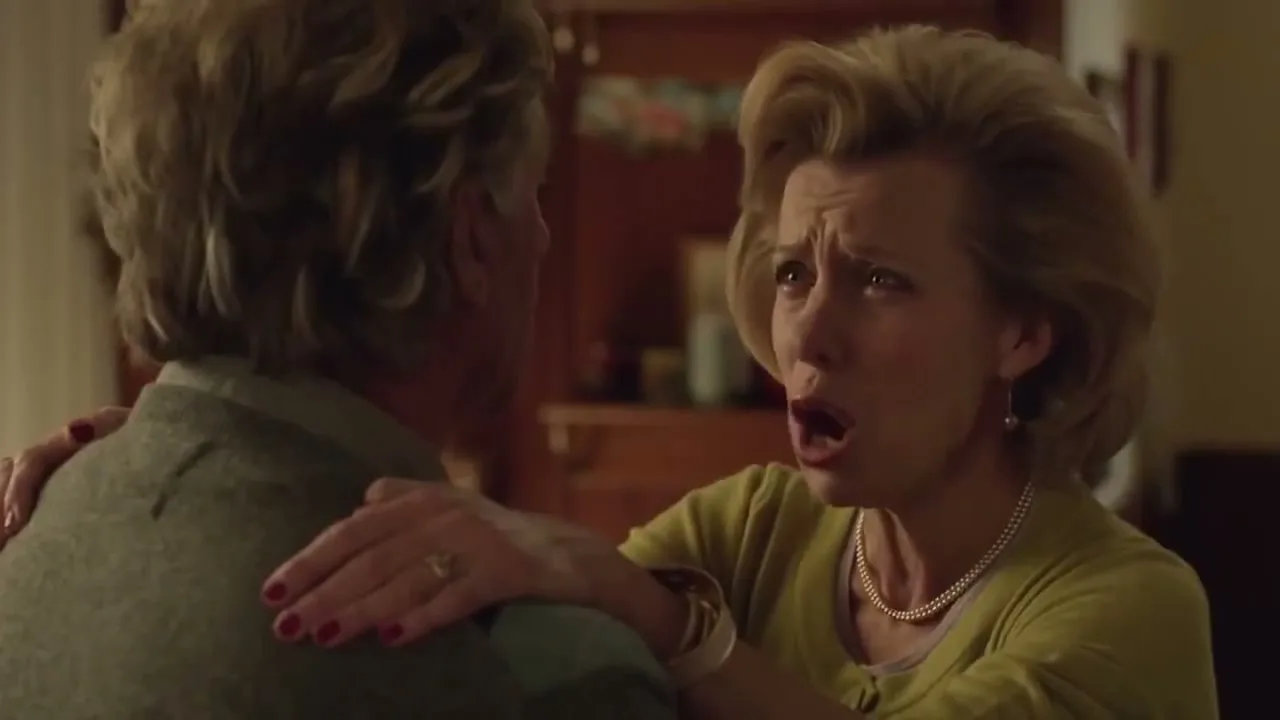 A blonde woman in a pearl necklace cries hysterically while holding onto a man's shoulders in a dimly lit living room.