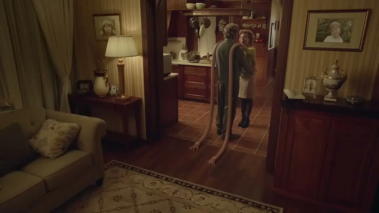 A middle-aged couple embraces in a cozy home, as the man's arms comically extend and slither across the floor, revealing a surreal transformation.