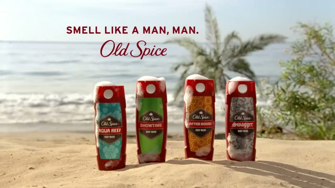 Four Old Spice body wash bottles, each topped with foam, stand in a row on a sandy beach with the ocean and a palm tree in the background.
