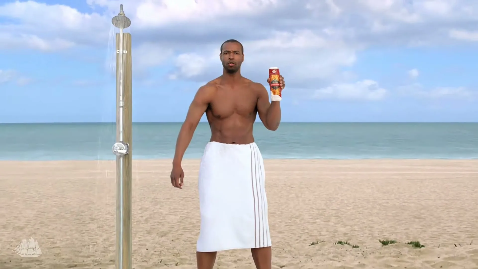 A shirtless man stands on a beach, holding a bottle of Old Spice body wash, then transitions to a lake with mountains and a cake.