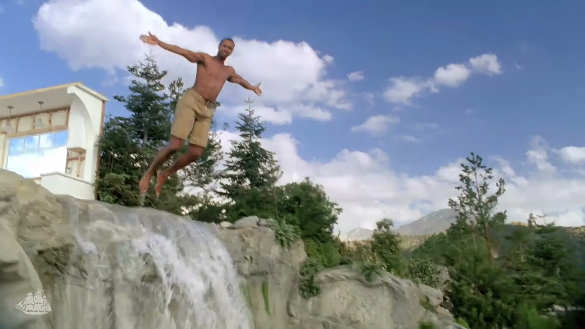 A shirtless man in khaki shorts jumps off a waterfall, arms outstretched, against a blue sky with clouds and mountains in the background.