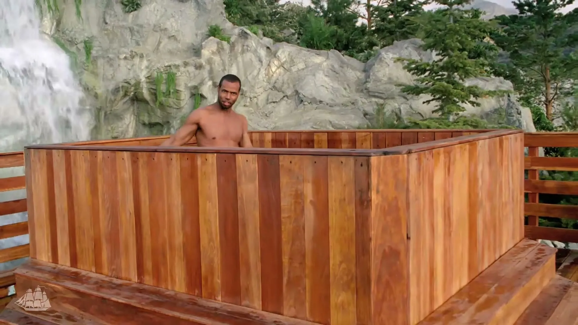 A shirtless man in his 30s stands in a wooden hot tub, splashing water, with a waterfall and rocky landscape in the background.