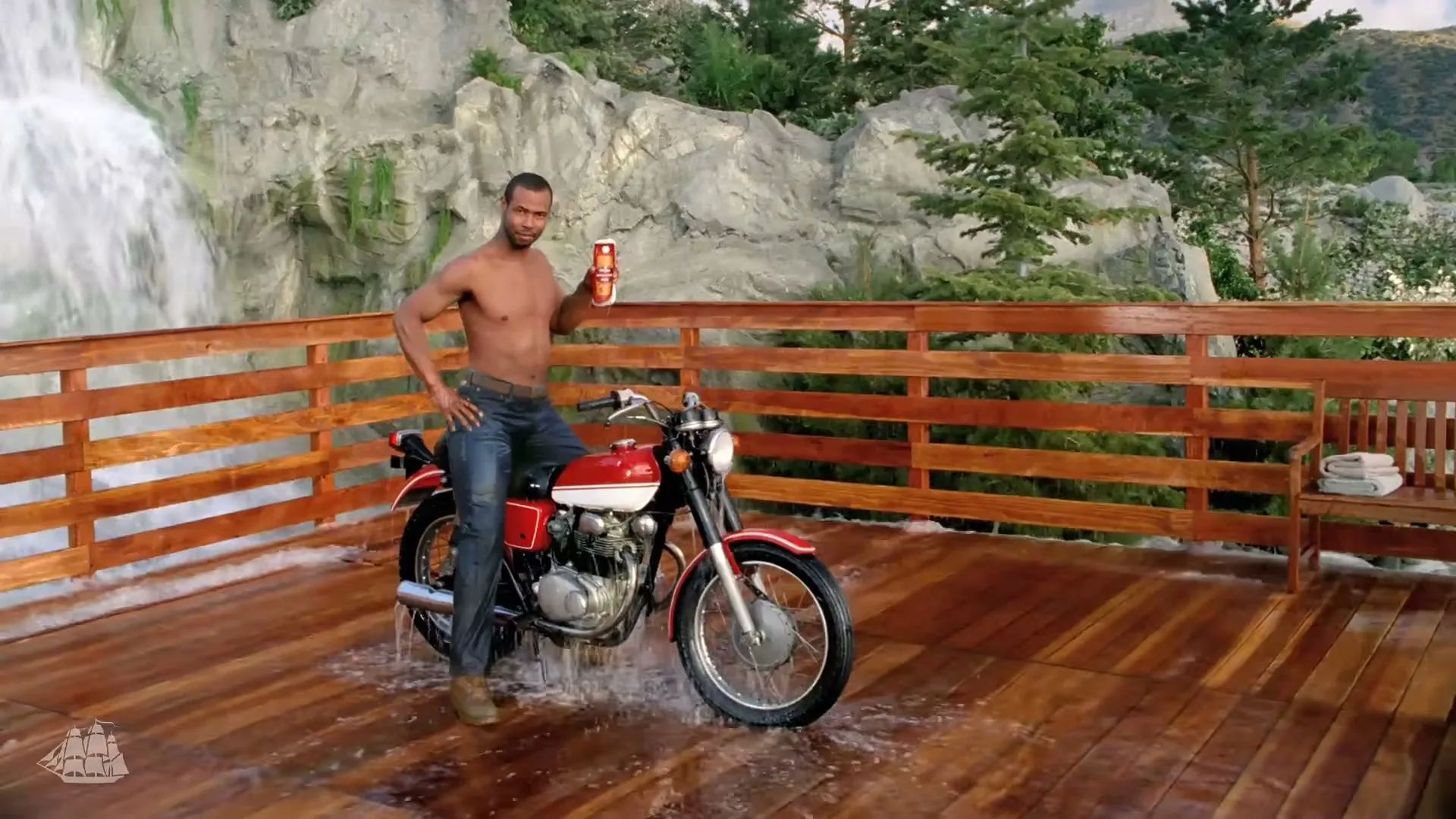 A shirtless man sits on a motorcycle on a wooden deck, holding an Old Spice product, as water recedes around him with a waterfall in the background.