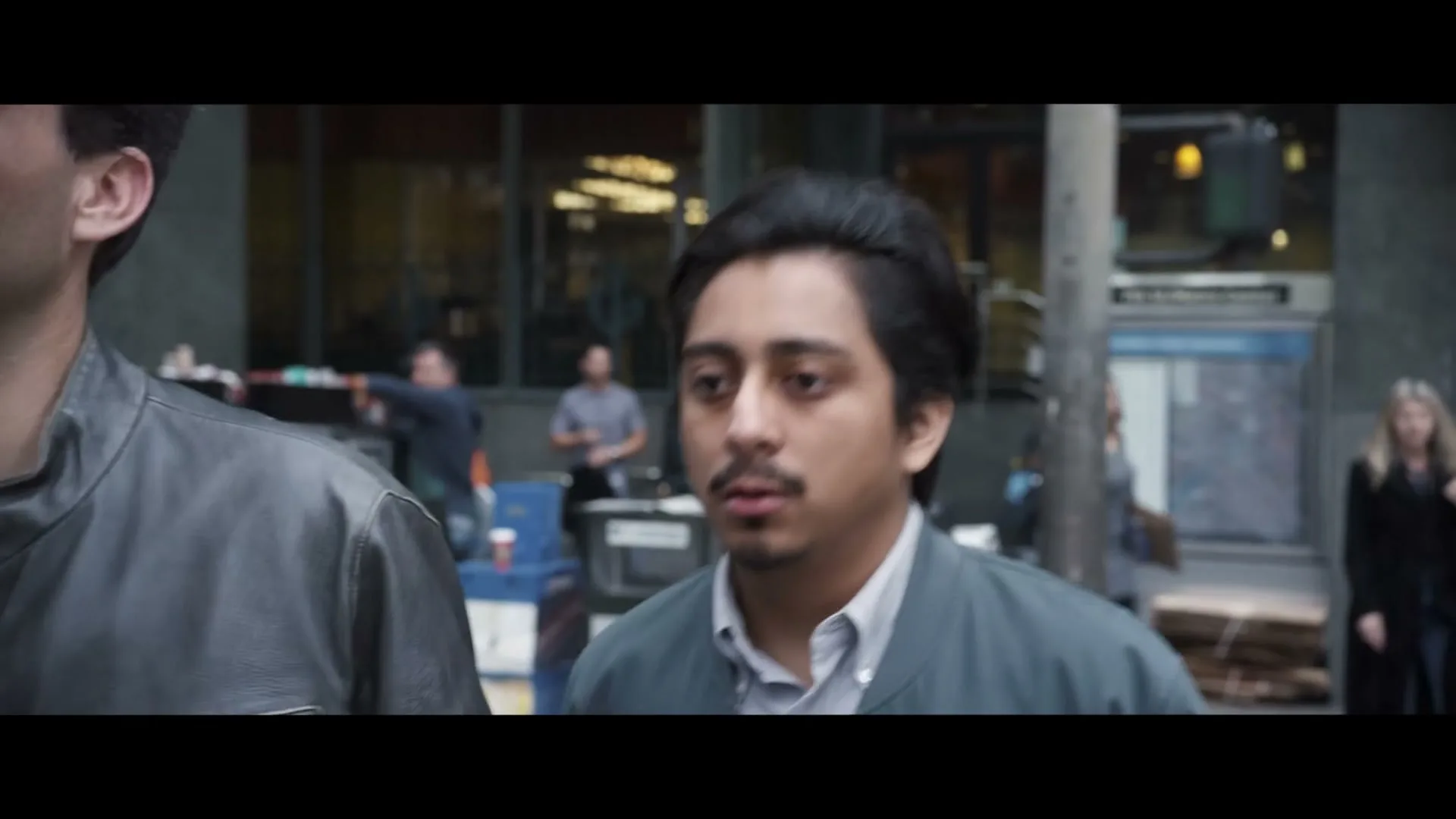 A young man with dark hair and a mustache, wearing a blue jacket, walks through a busy city street, looking around with a slightly concerned expression.