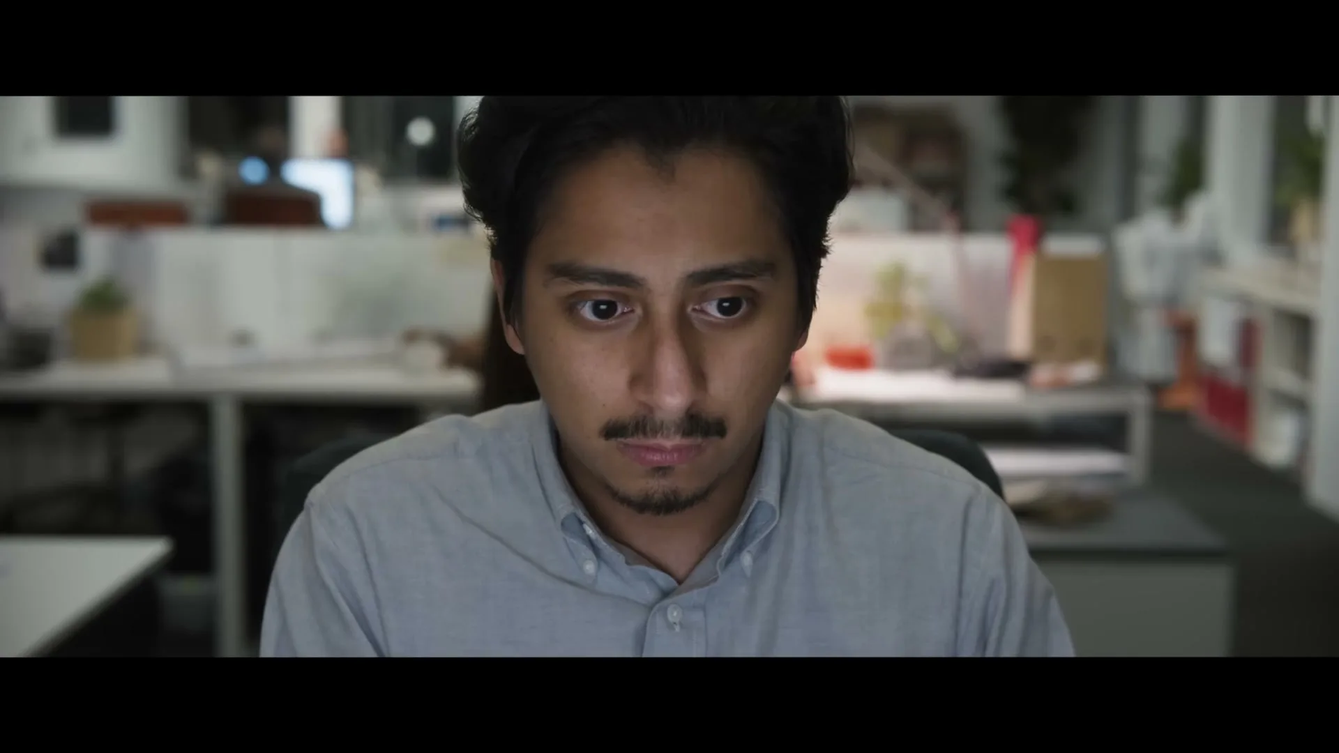 A young man with dark hair and a mustache sits at a desk in a dimly lit office, looking intently at a screen.