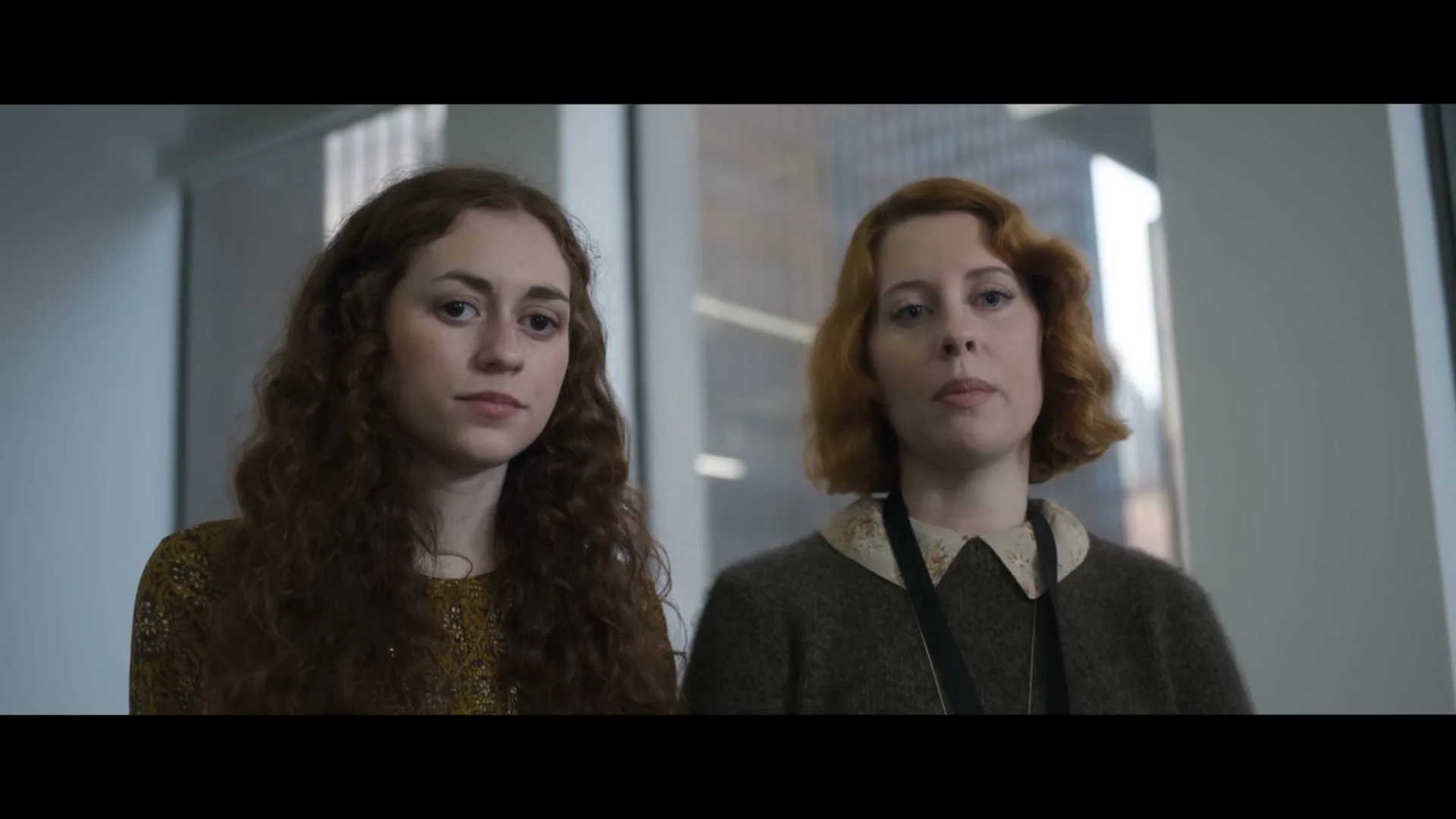Two young women, one with curly brown hair and the other with red hair, stand side-by-side in an office building, looking forward with neutral expressions.