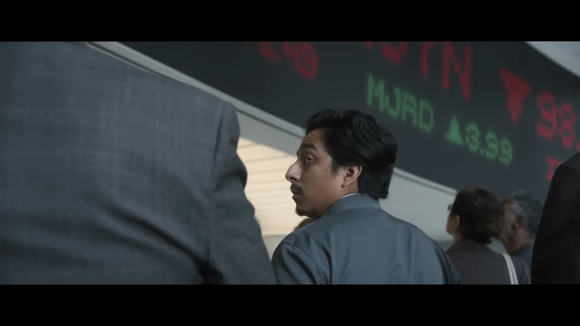 A man with dark hair and a grey jacket looks up at a stock ticker with red and green numbers, surrounded by other people in a busy financial setting.