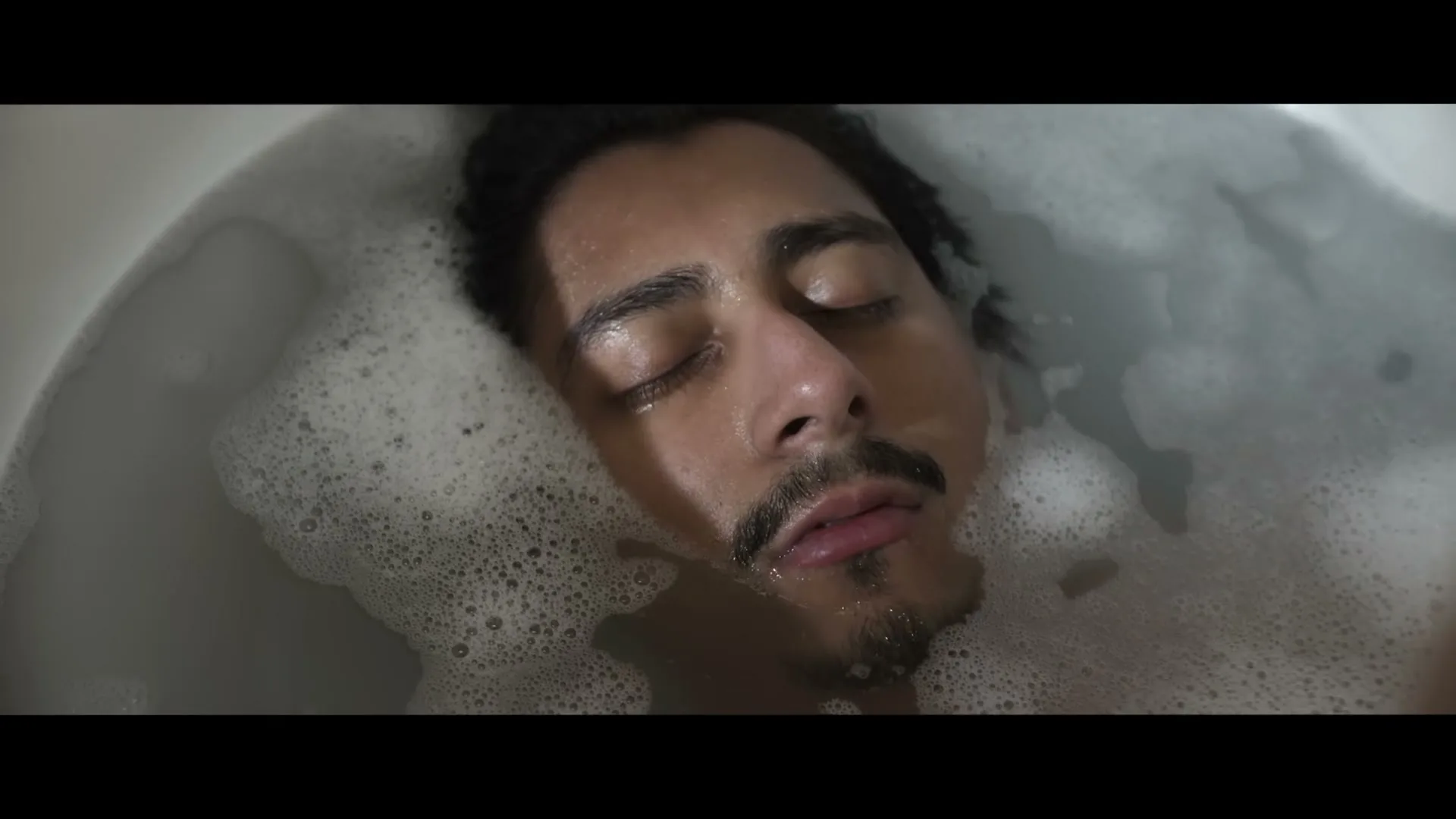 A young man with a mustache and closed eyes lies submerged in a bathtub filled with bubbles, his face illuminated by soft light.