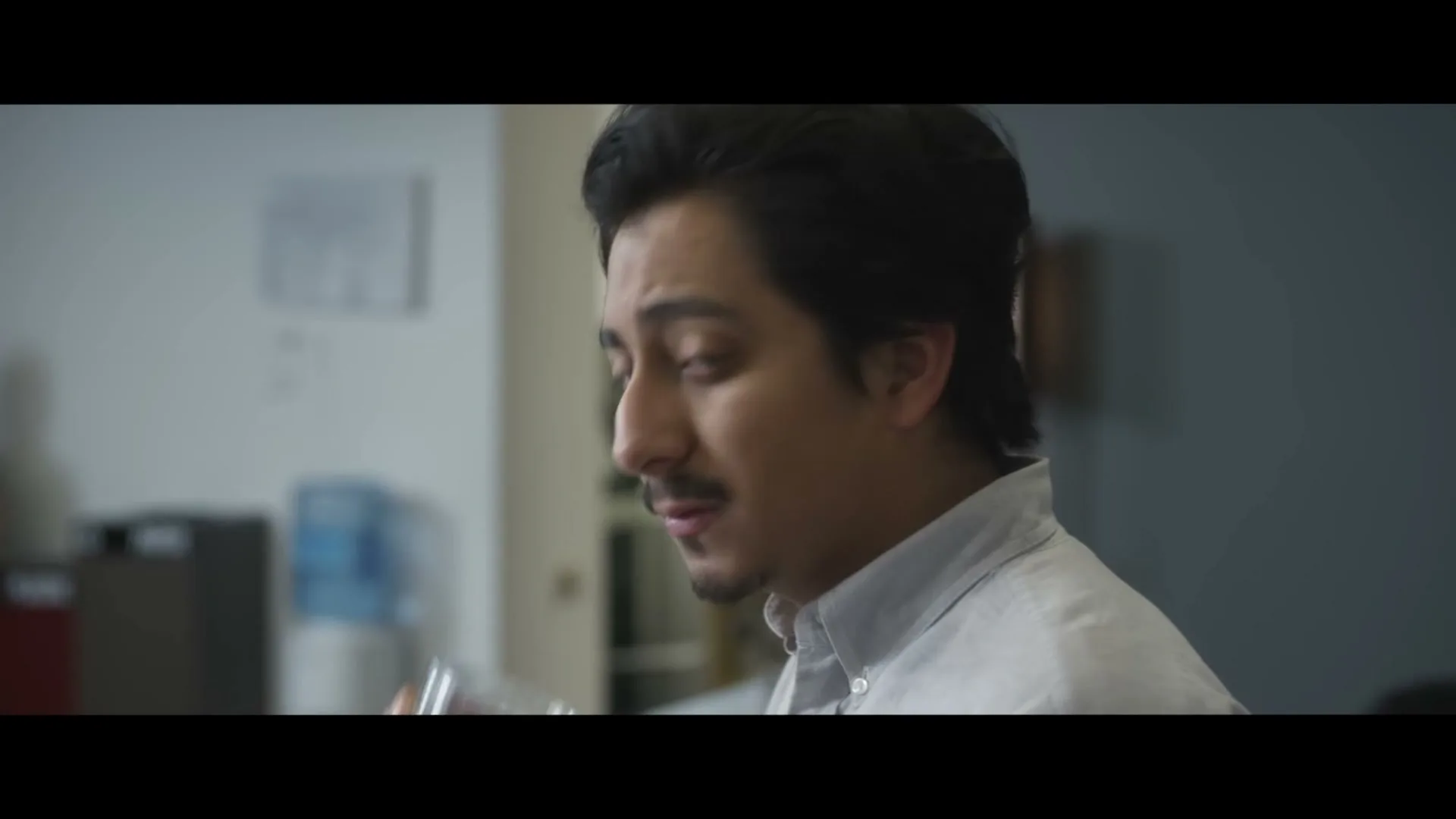 A young man with dark hair and a mustache drinks water from a glass, then looks towards the camera with a concerned expression in an office setting.