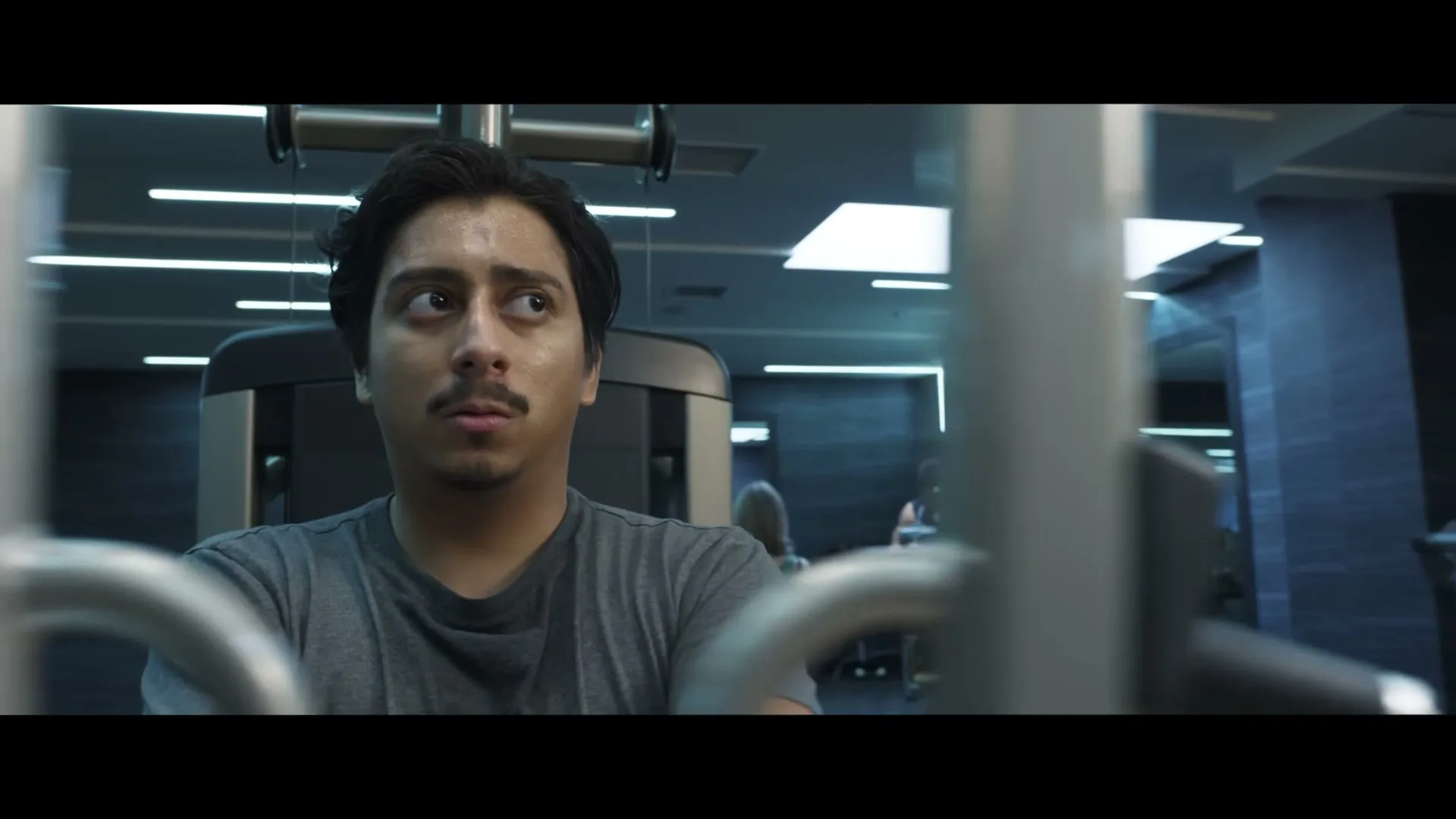 A man with a mustache and dark hair, wearing a grey t-shirt, looks around with a worried expression while working out in a gym.