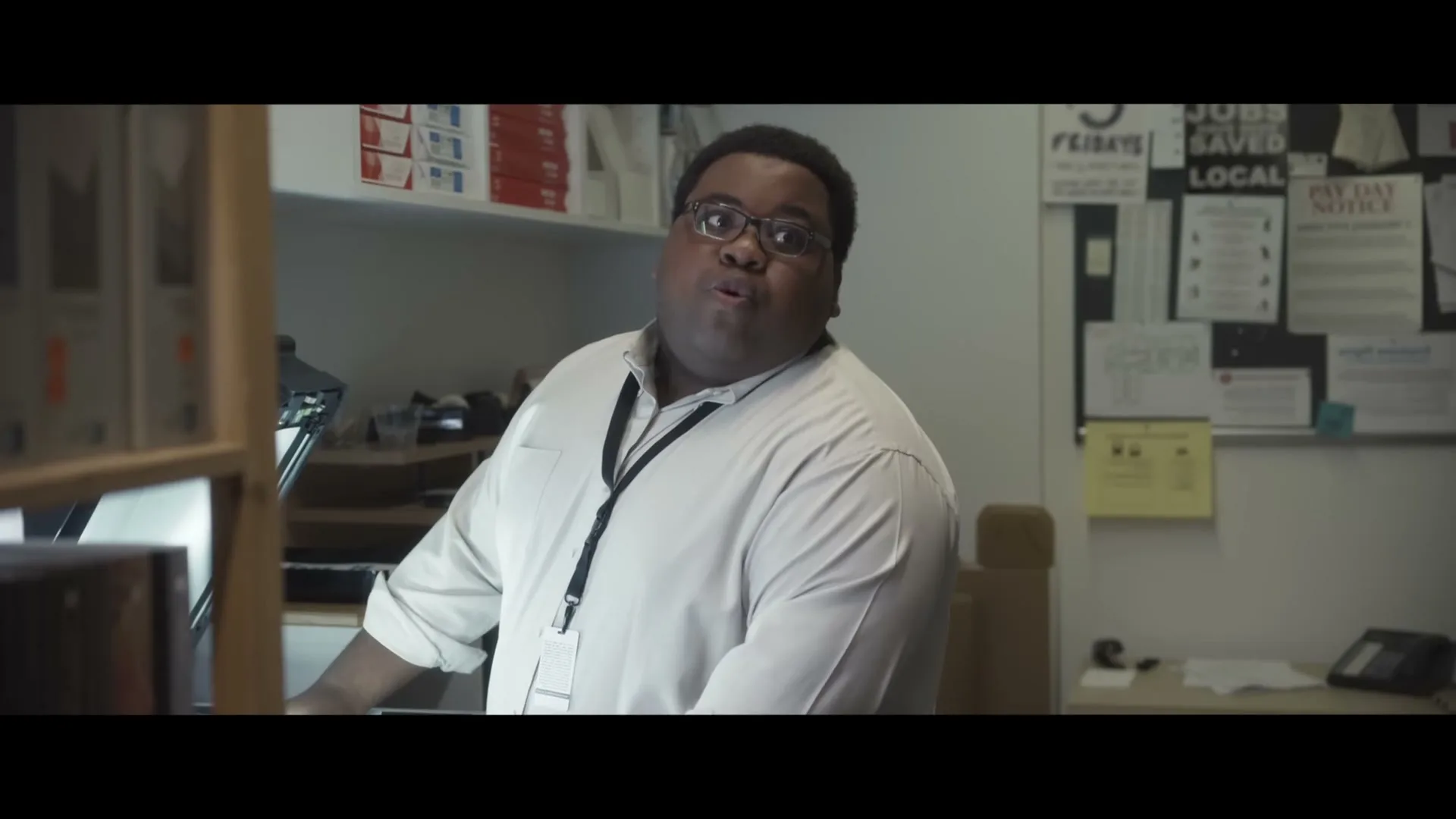 A Black man in his 30s, wearing glasses and a white shirt, looks surprised and speaks in an office setting.
