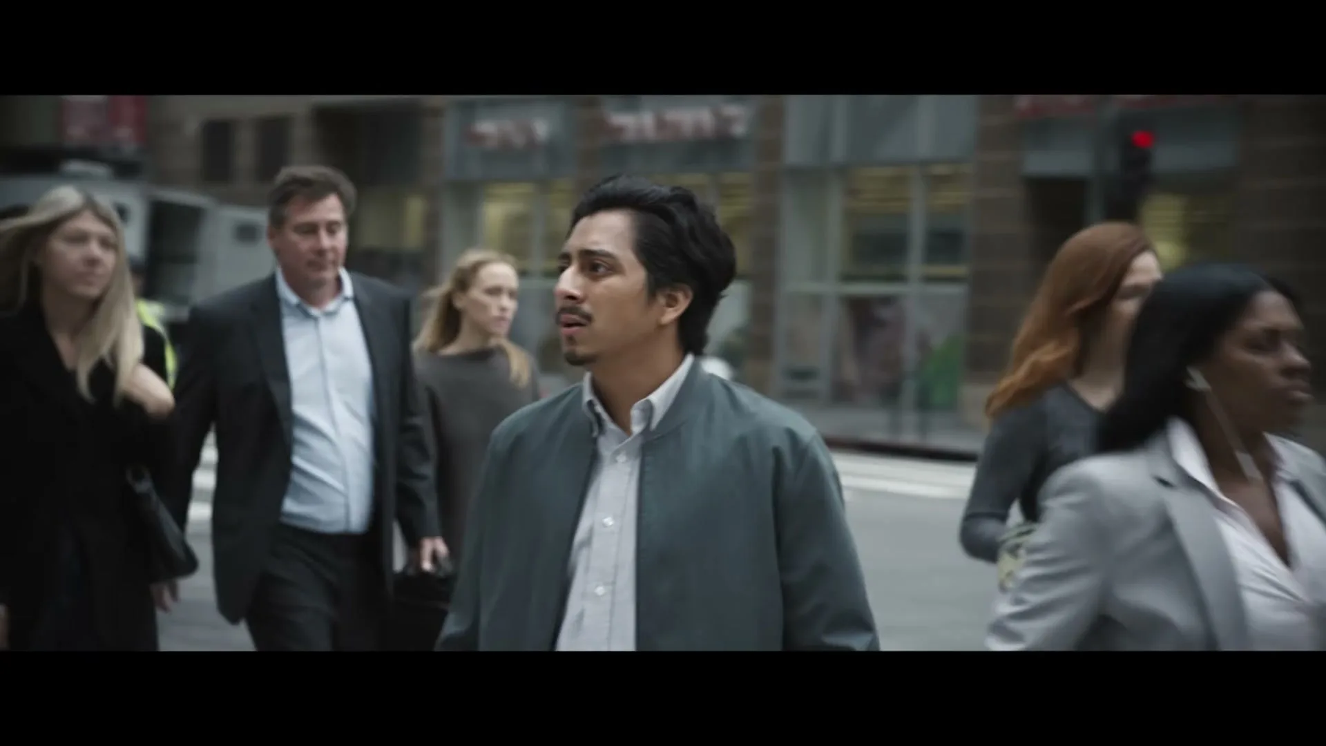 A young man with dark hair and a mustache looks around nervously while walking through a busy city street with other pedestrians.