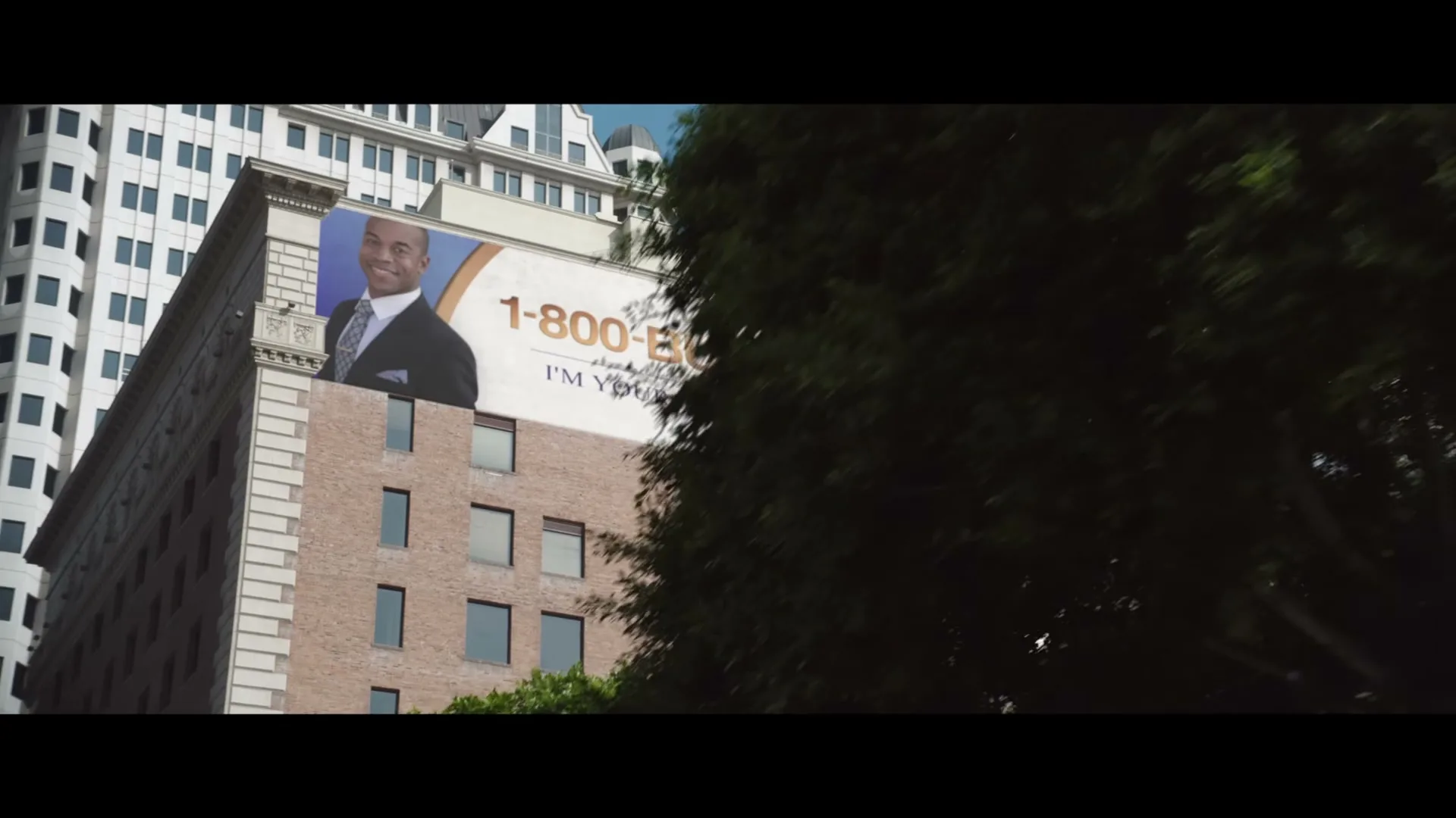 A man with dark hair and a mustache runs past a building with a large billboard that reads "1-800-BUY-NOW I'M YOUR MONEY GUY."