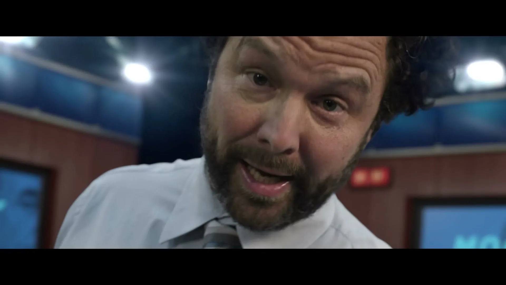 A disheveled male news anchor with a beard and tie looks directly at the camera, speaking in a news studio.
