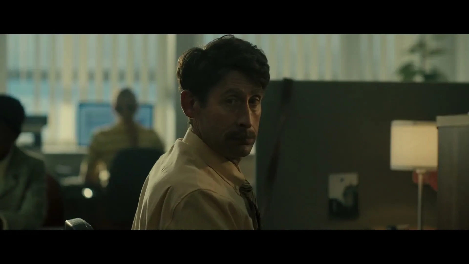 A man with a mustache in a beige shirt and tie turns his head to look over his shoulder in a dimly lit office.