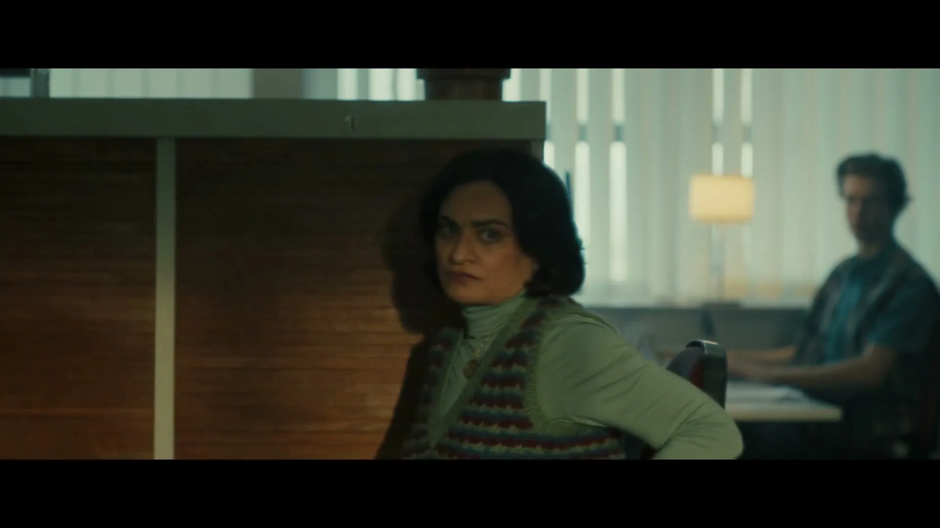 A woman in a green top and patterned vest looks around nervously in an office cubicle, while a man works at a desk in the background.