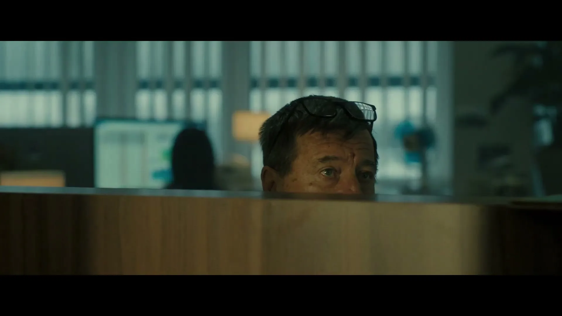 A man with glasses on his head peeks over a cubicle wall, looking around an office with a worried expression.