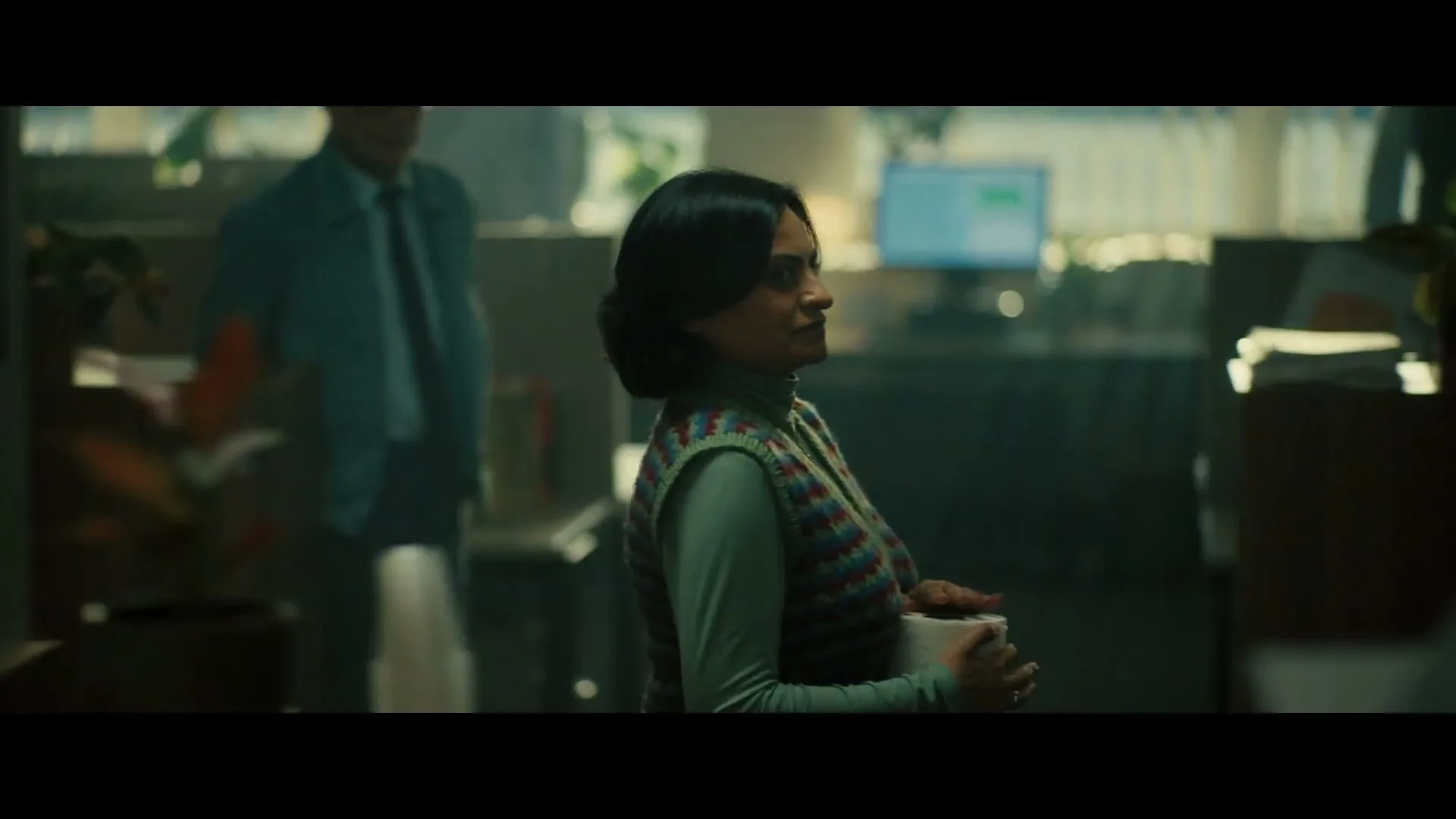 A woman in a patterned vest and turtleneck holds a mug and looks around an office, as a man walks past in the background.