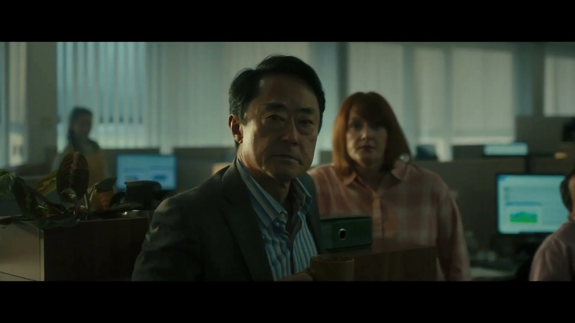 A middle-aged Asian man in a suit holds a mug and looks directly at the camera in a dimly lit office, with other colleagues in the background.