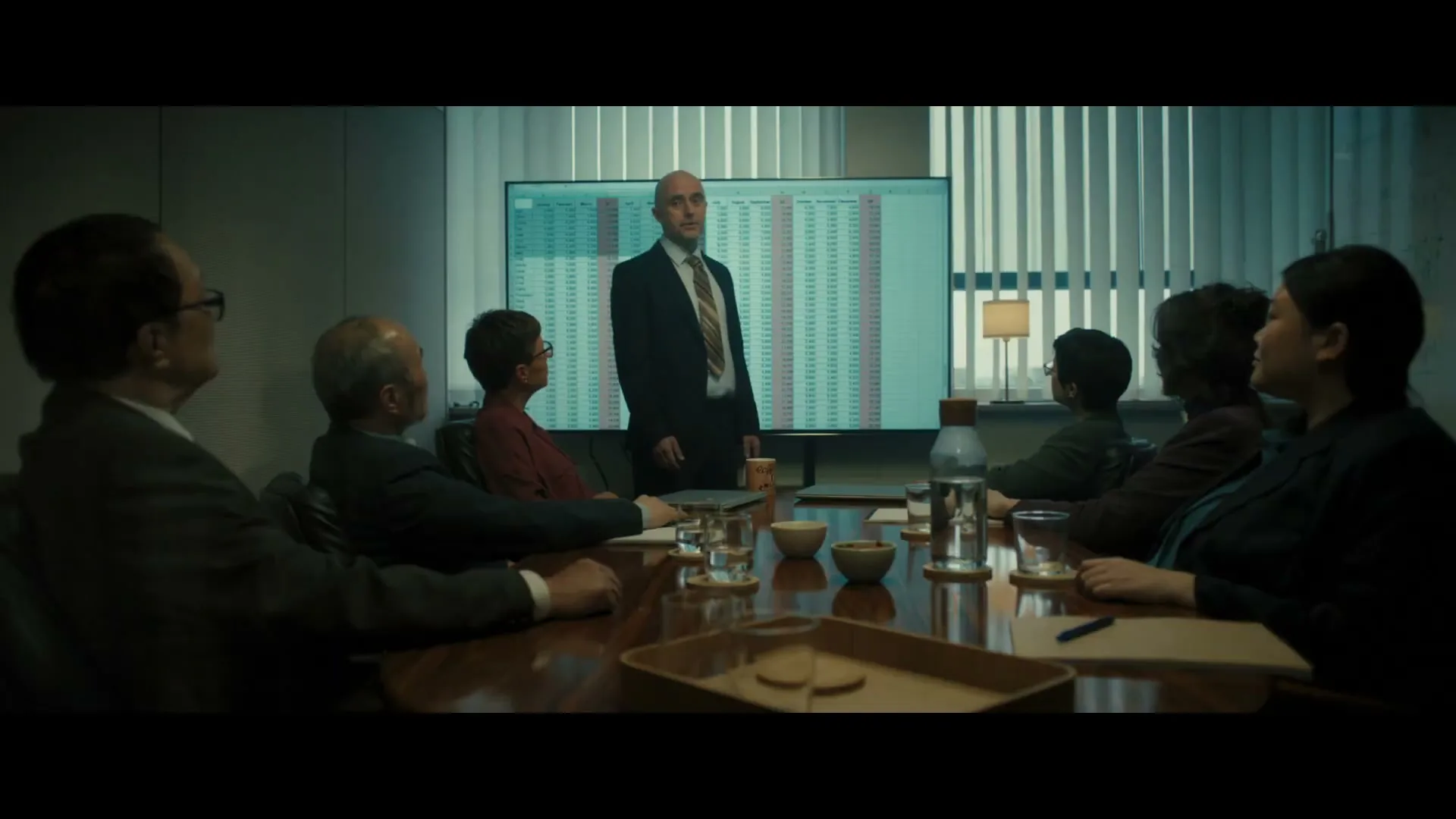 A bald man in a suit presents a spreadsheet on a large screen to a diverse group of colleagues sitting around a conference table in an office.