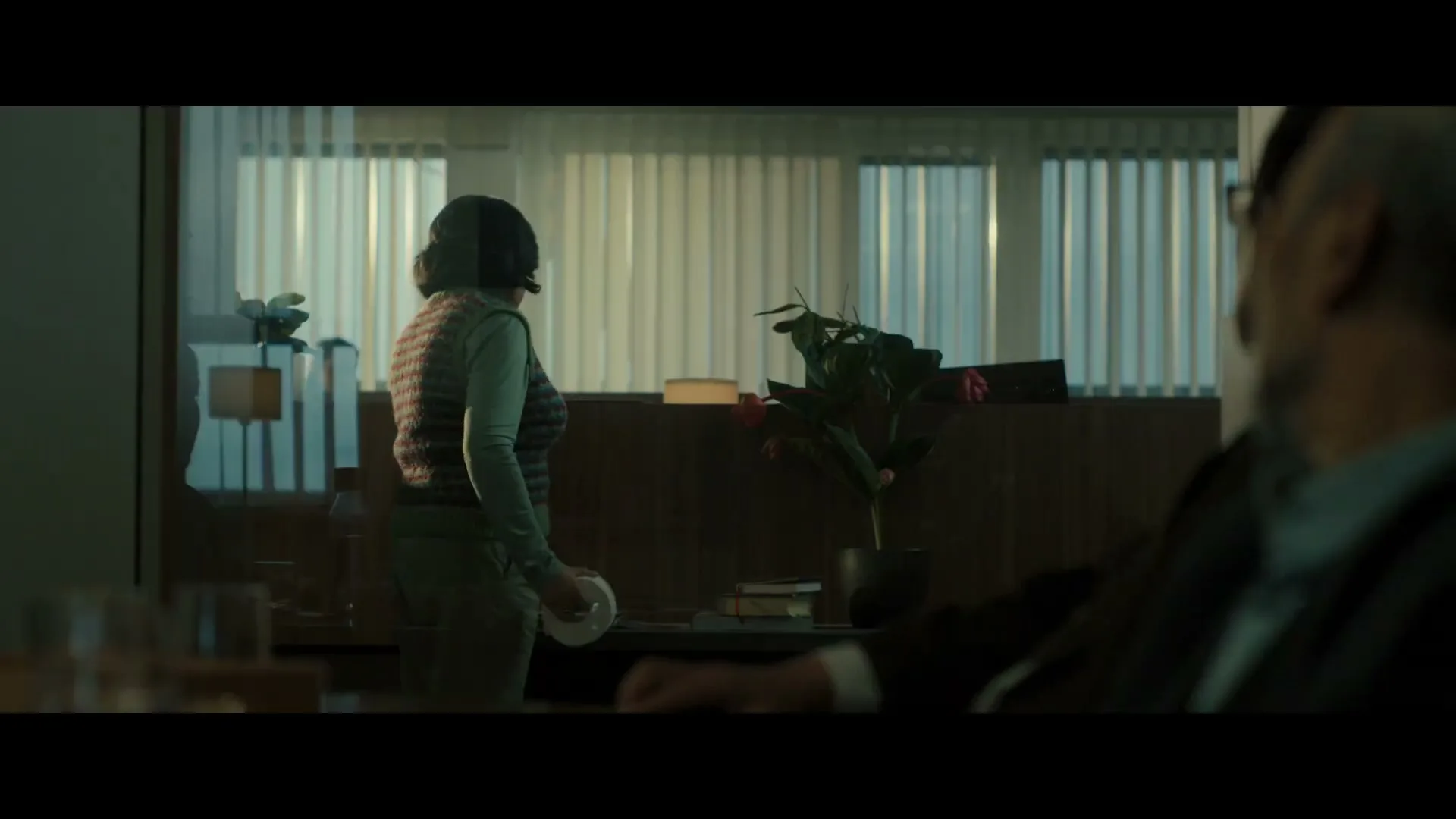 A woman in a sweater vest walks past a desk, holding a roll of toilet paper, while a man sits in the foreground.