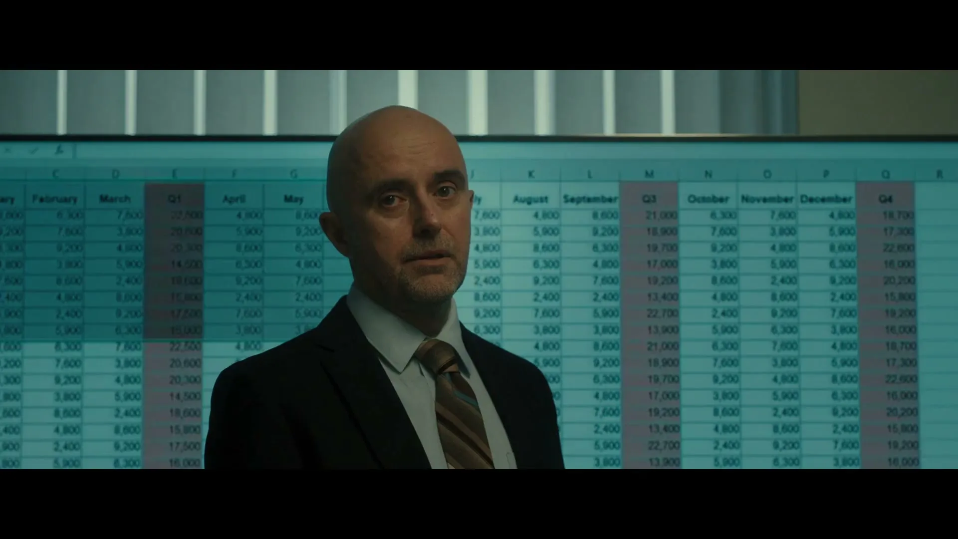 A bald man in a suit and tie looks directly at the camera with a serious expression in an office with a large spreadsheet on a screen behind him.