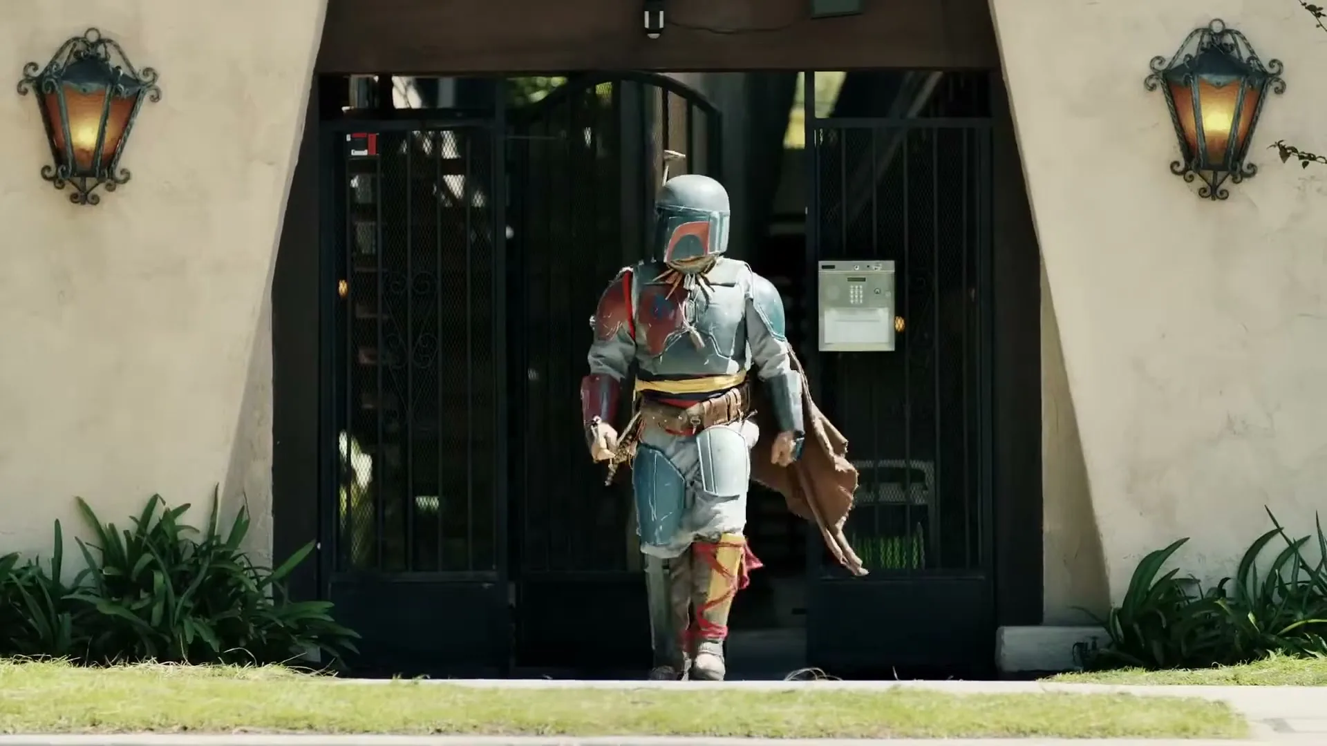 A person dressed as Boba Fett from Star Wars walks out of a house gate onto a suburban sidewalk on a sunny day.