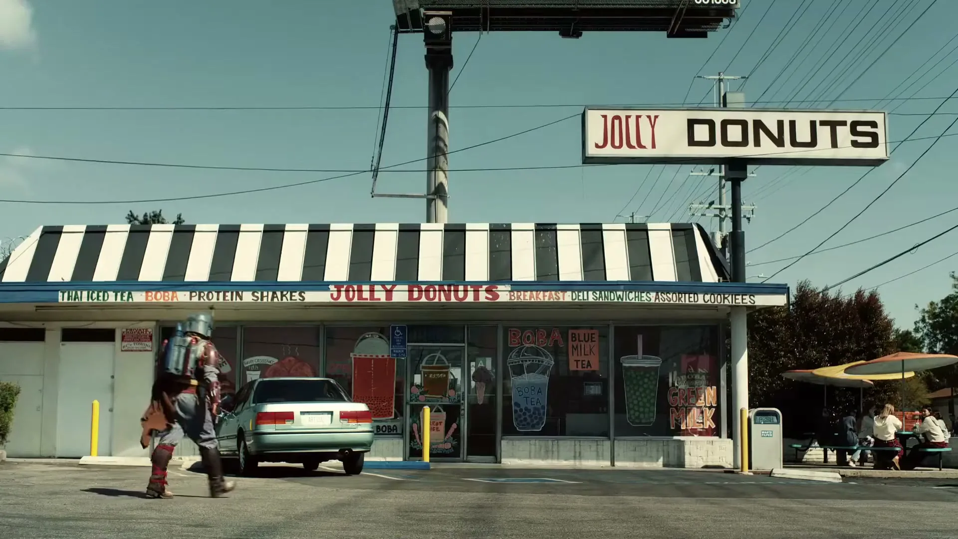 A person in a full Mandalorian costume walks past a retro donut shop with a striped awning and a parked car in front of it on a sunny day.