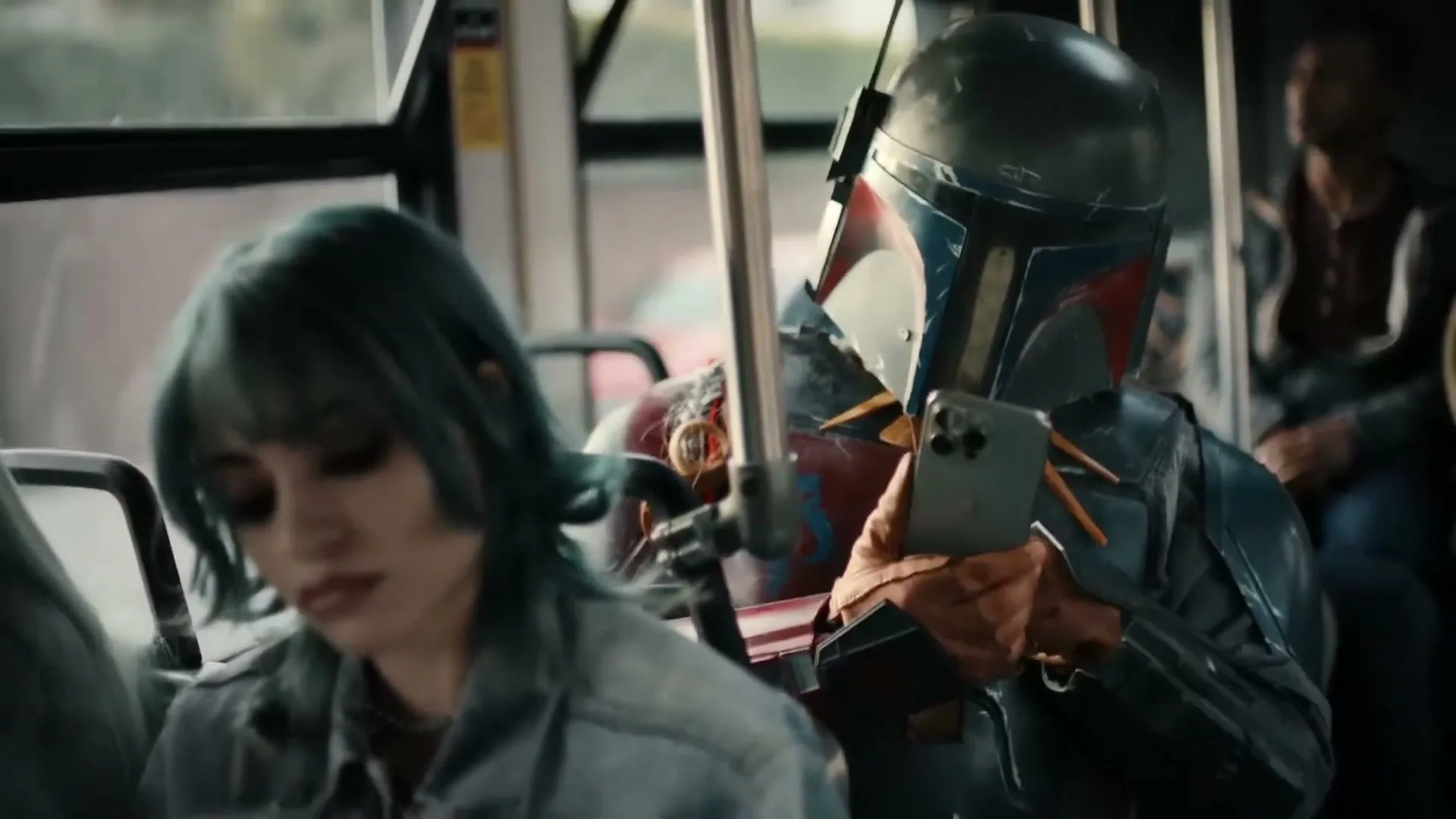 A person in a Boba Fett costume holds an iPhone on a bus, while a young woman with blue hair sits in the foreground.