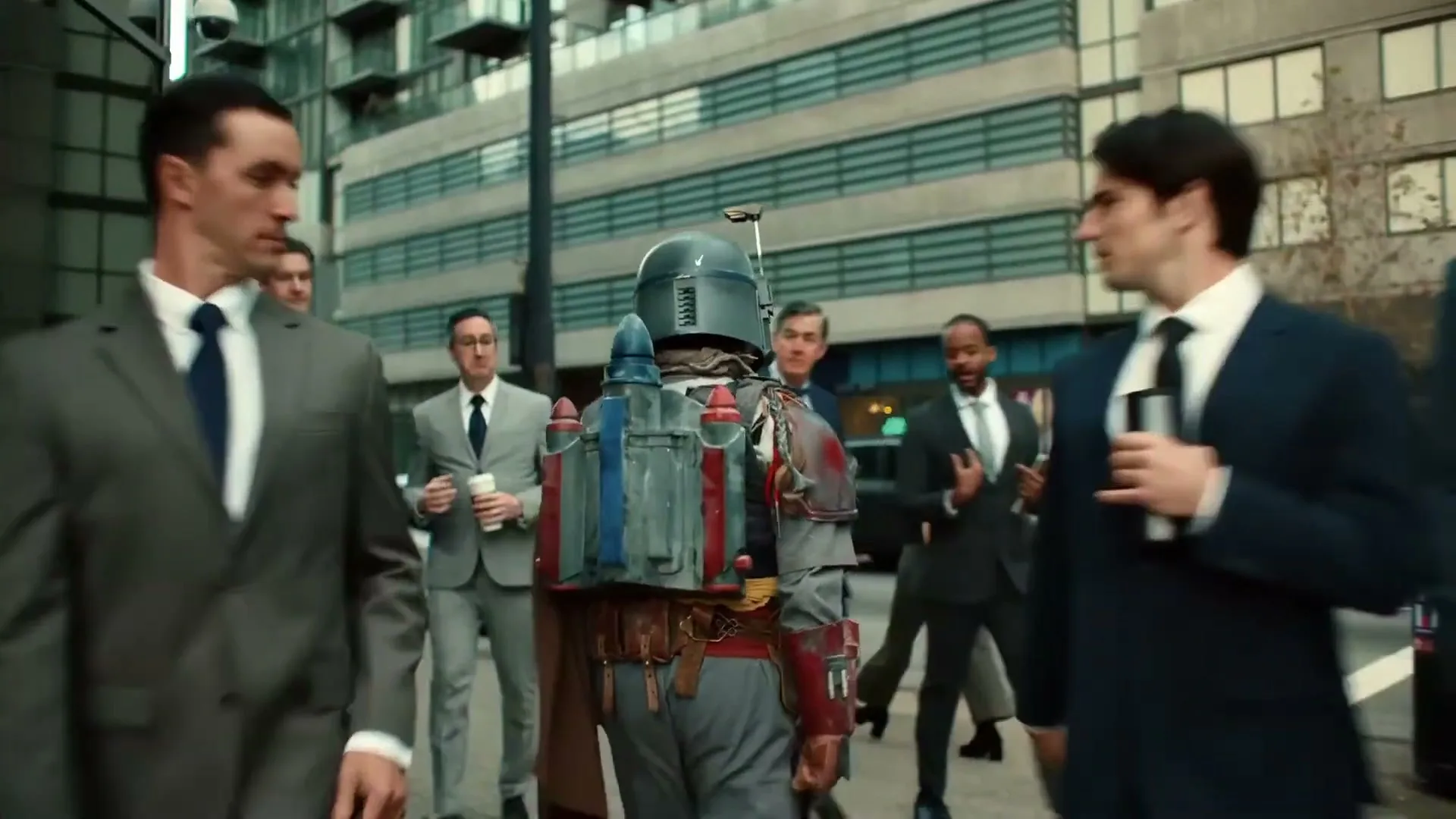 A person dressed as Boba Fett walks down a city street, surrounded by business people in suits, with modern buildings in the background.